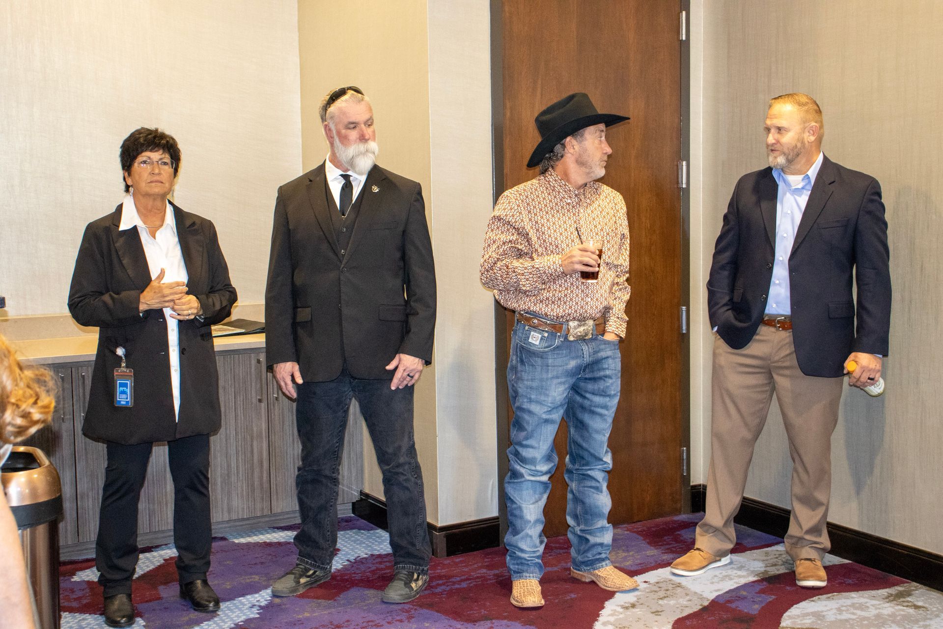 Four people stand in an indoor setting, three in professional attire and one wearing a cowboy hat and patterned shirt.