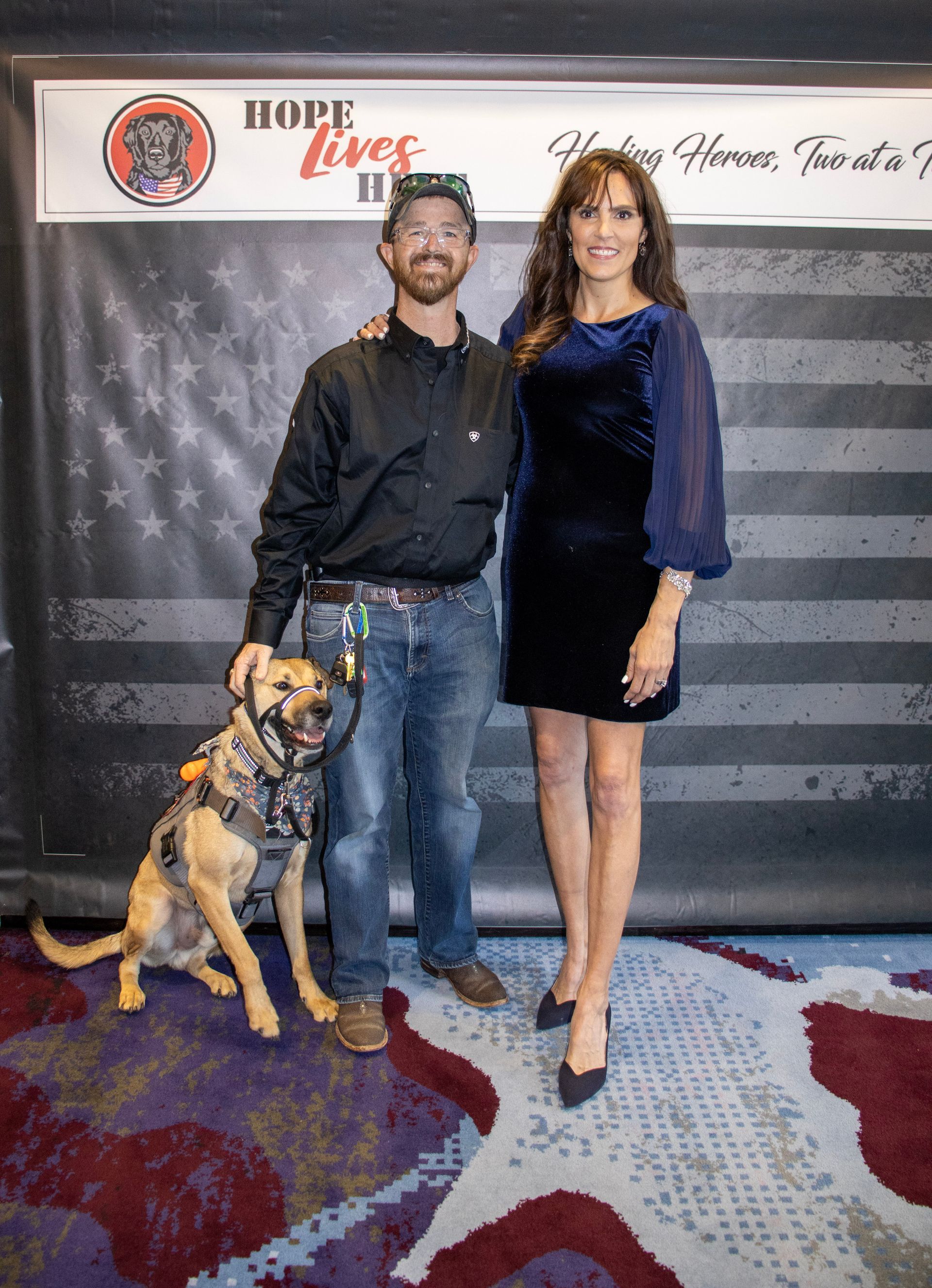 A person wearing a cap, black shirt, and jeans stands with a service dog next to a person in a navy velvet dress.