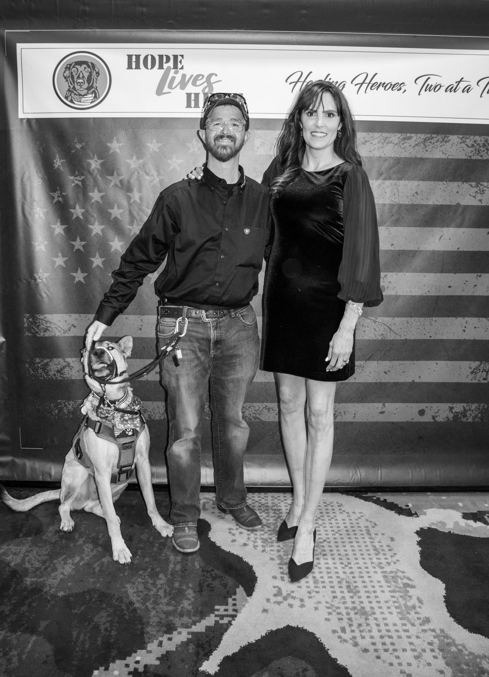 A man in a button-down shirt and jeans stands with a service dog next to a woman in a dress against a flag backdrop.