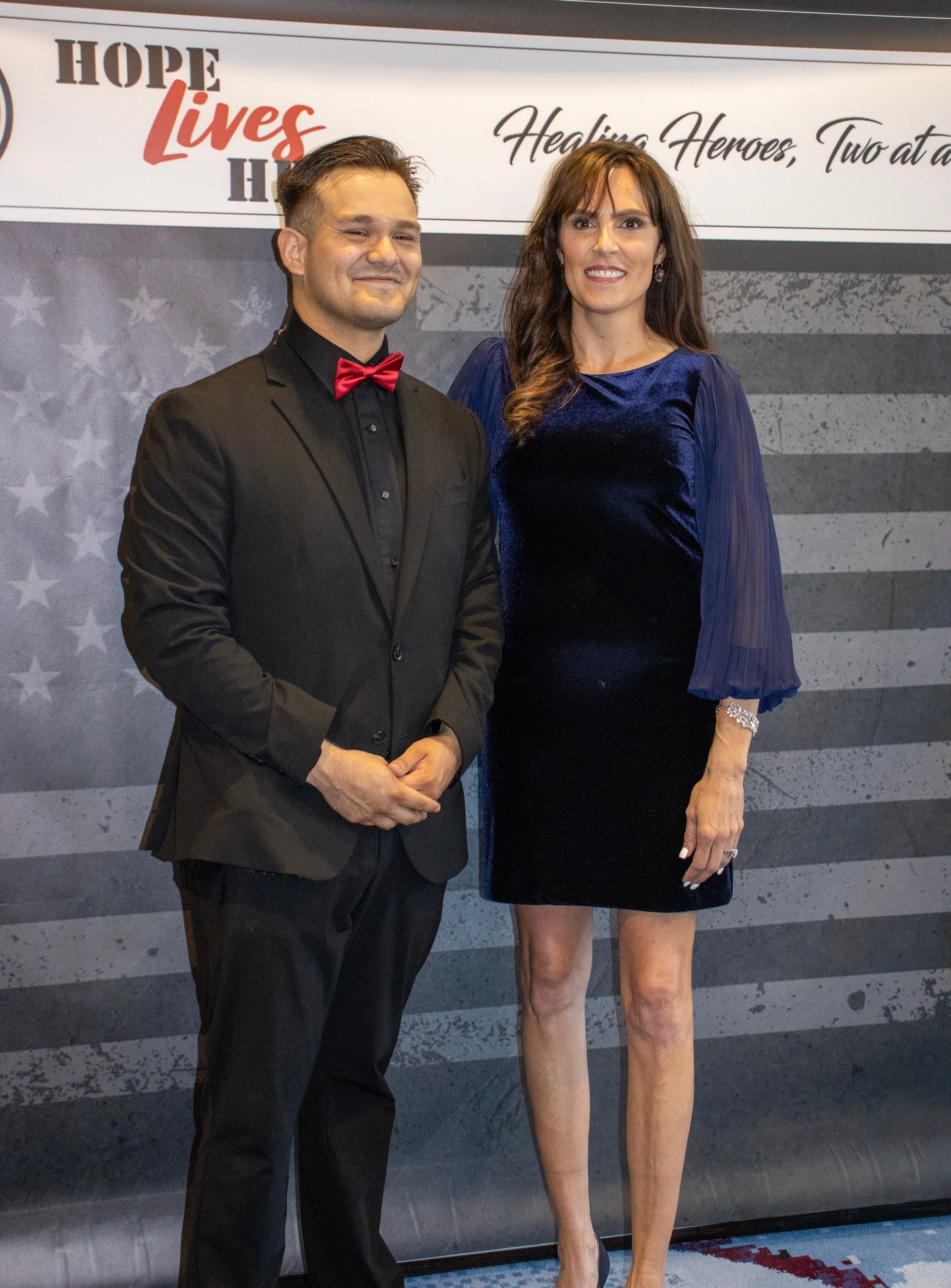 A person in a suit with a red bow tie and a person in a navy velvet dress stand before a US flag backdrop.