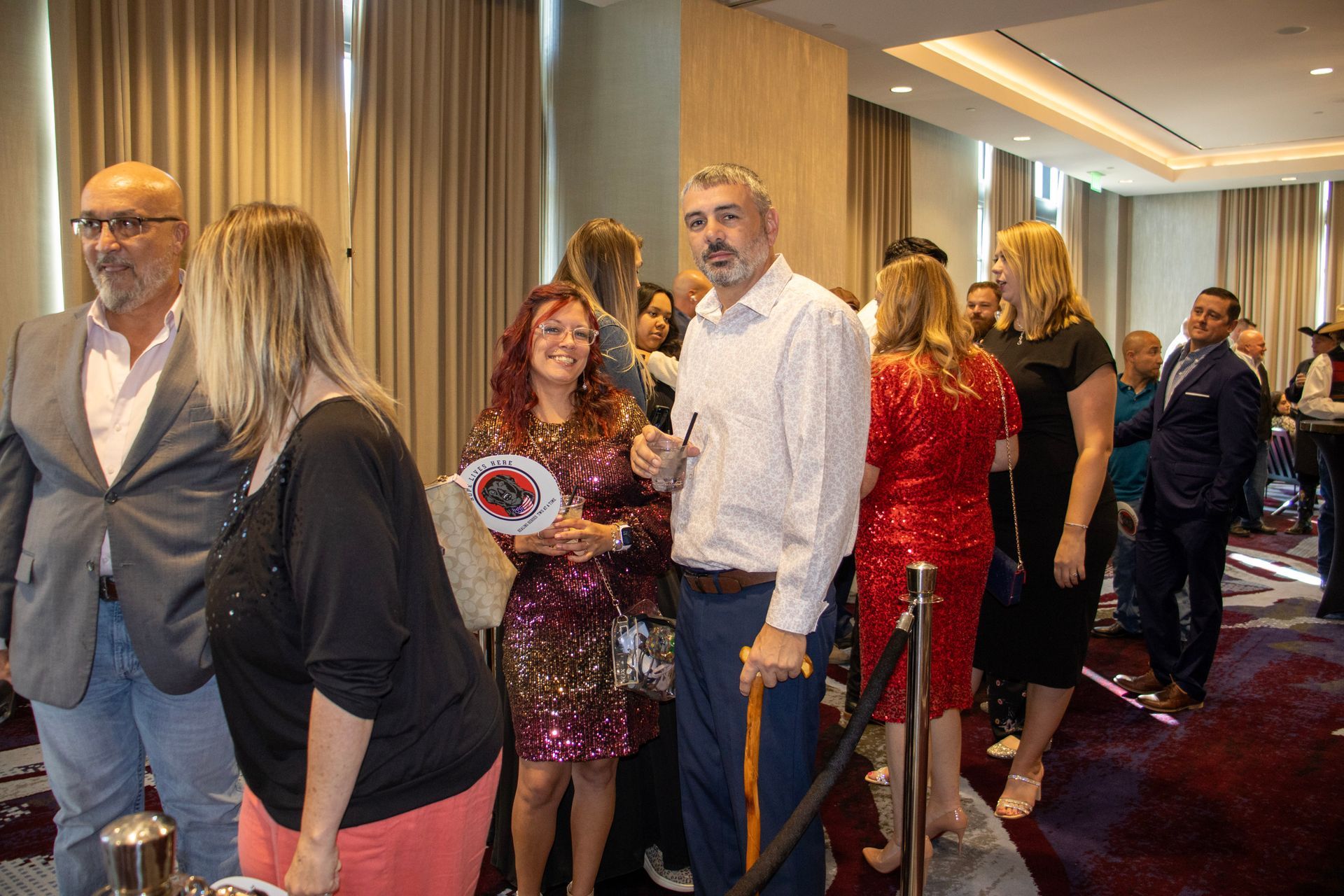 People at an indoor event, standing in a line in a room with floor-to-ceiling curtains and patterned carpet.
