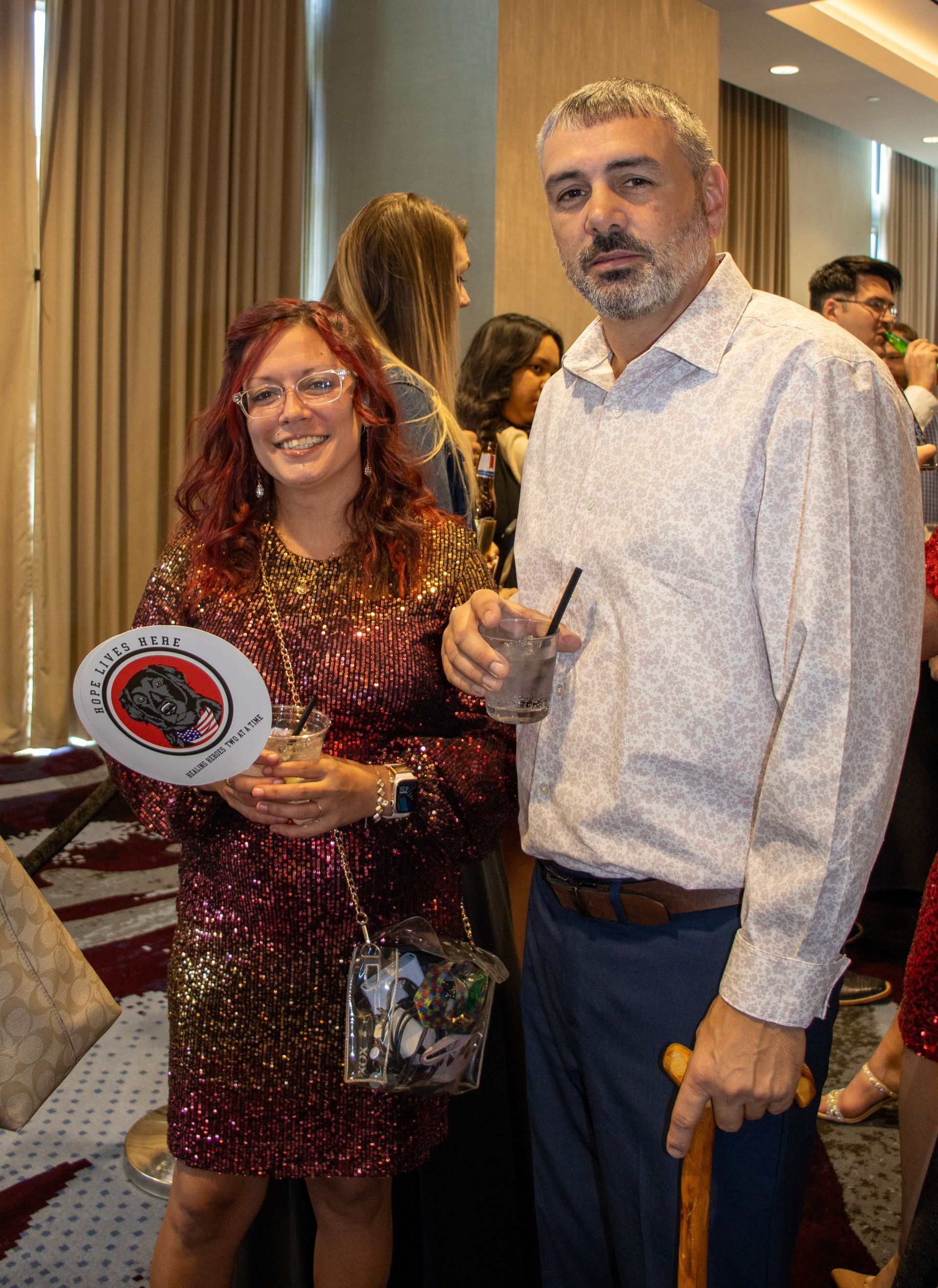 A smiling person in a red sequin dress and a man with a beard holding a drink and a wooden cane stand at an indoor event.