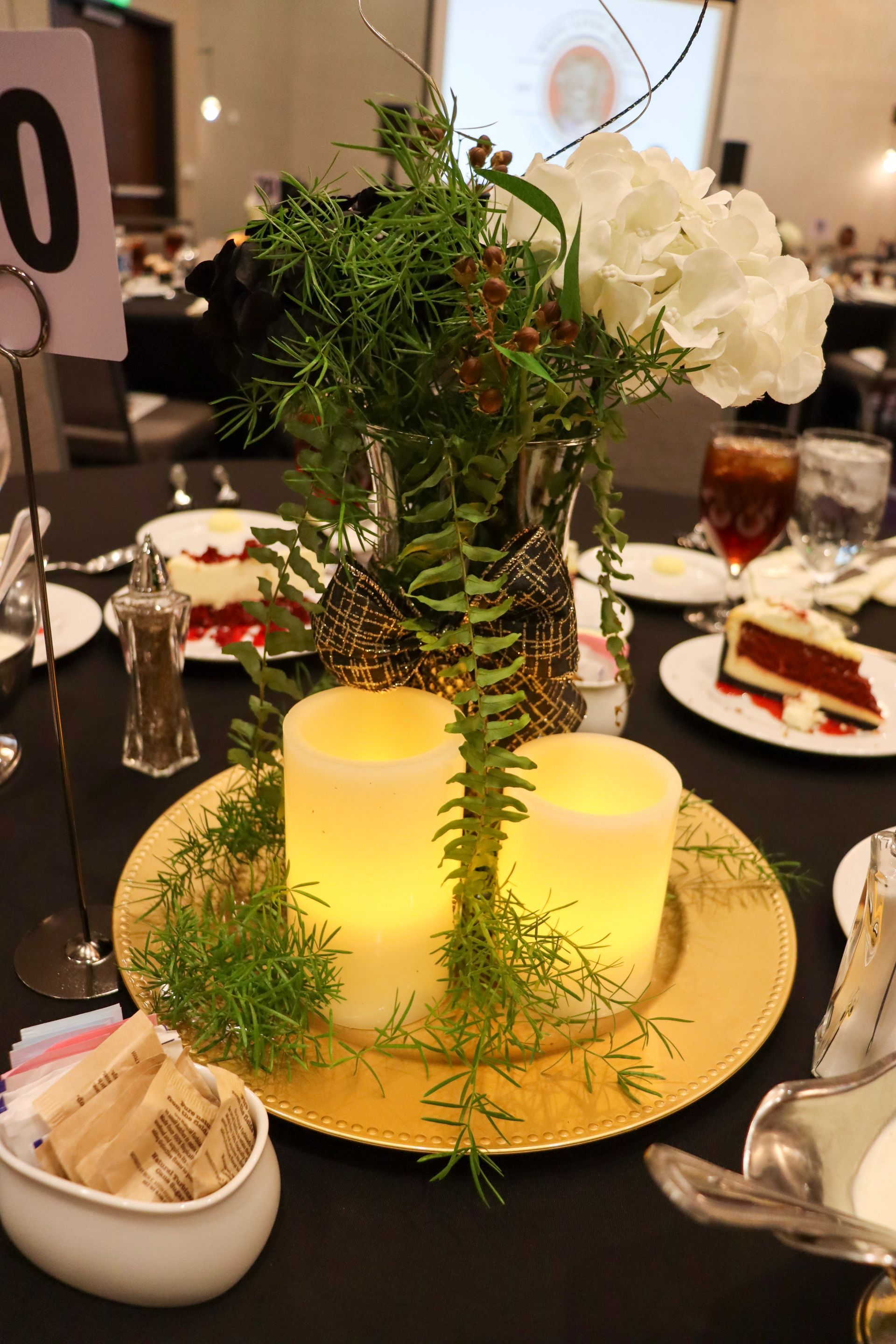A centerpiece with flowers, greenery, and two LED pillar candles on a gold tray, set on a table at a banquet event.