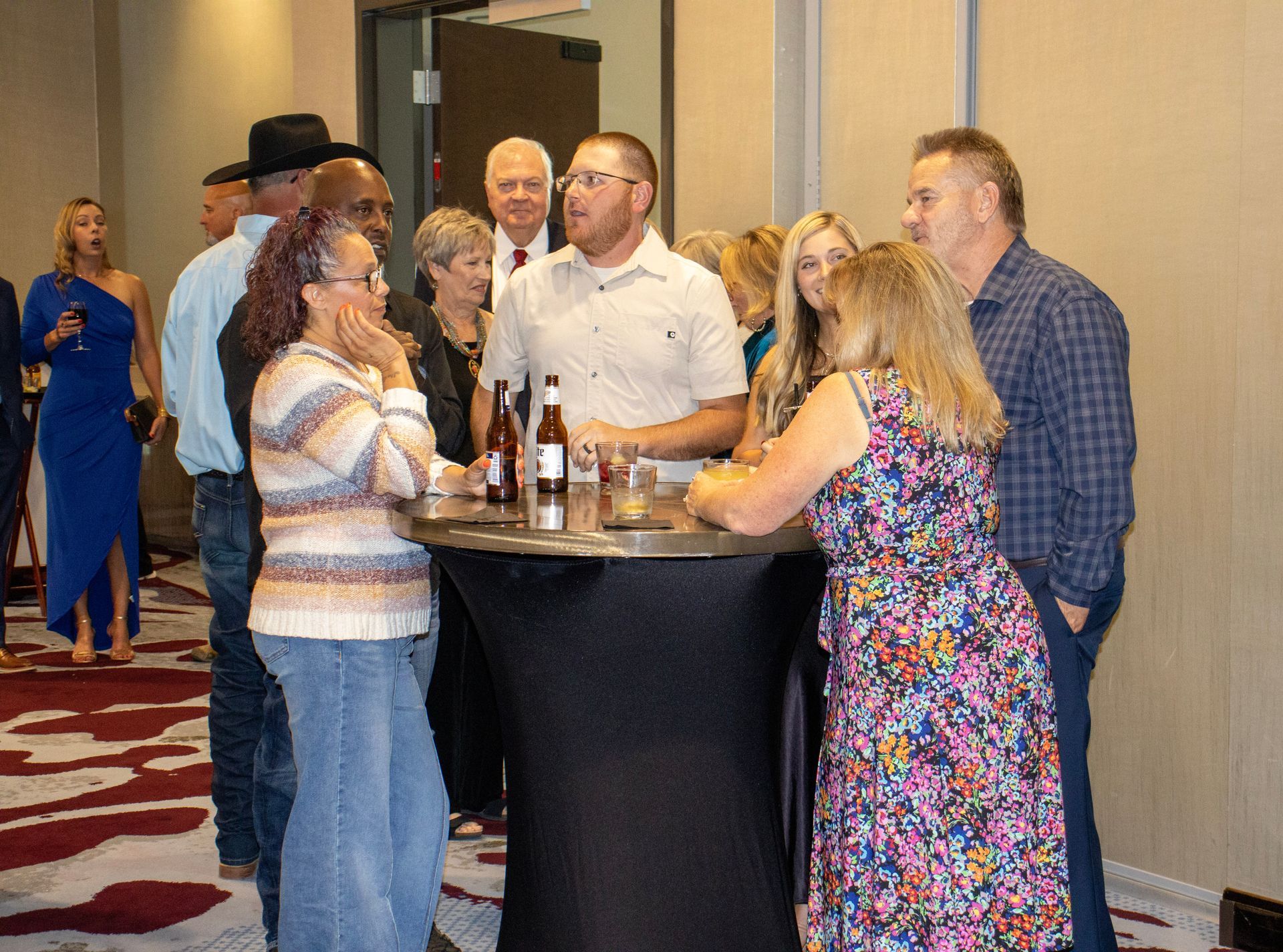 A group of people standing around a high cocktail table with drinks in an indoor event space with patterned carpeting.