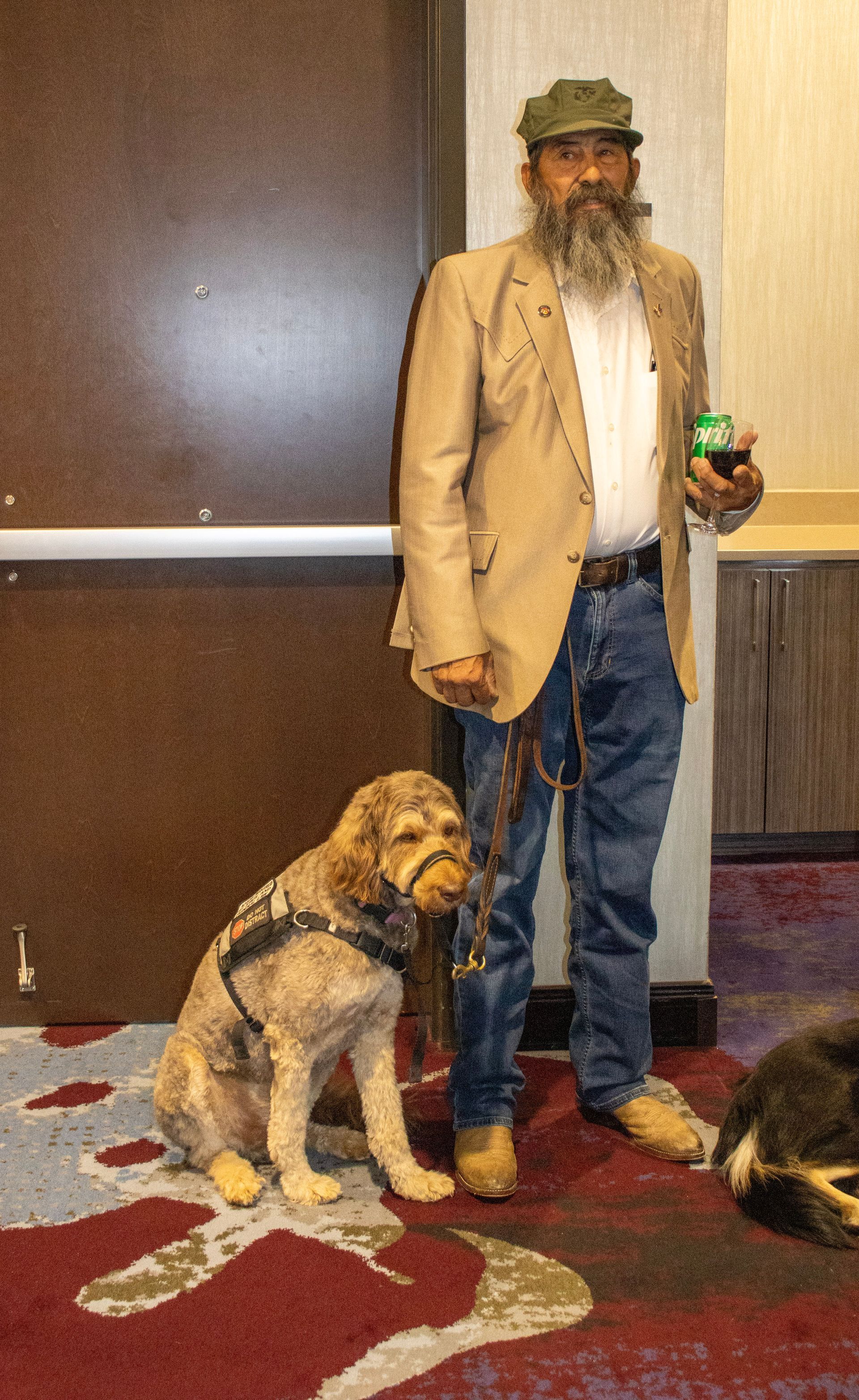 A bearded person in a tan jacket and cap stands on a patterned carpet next to a seated light-brown dog on a leash.