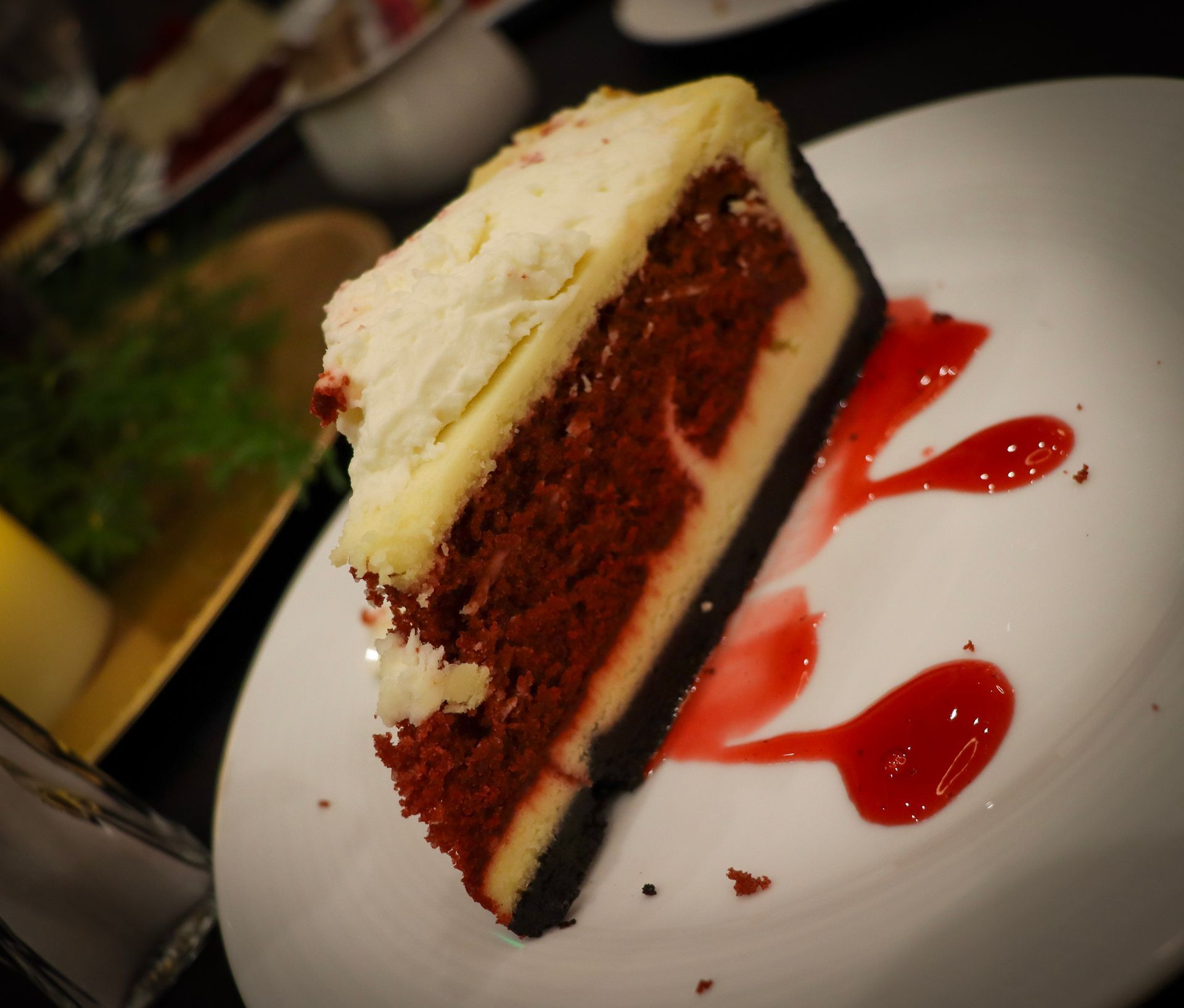 A slice of red velvet cheesecake on a white plate, decorated with dots of red berry coulis.