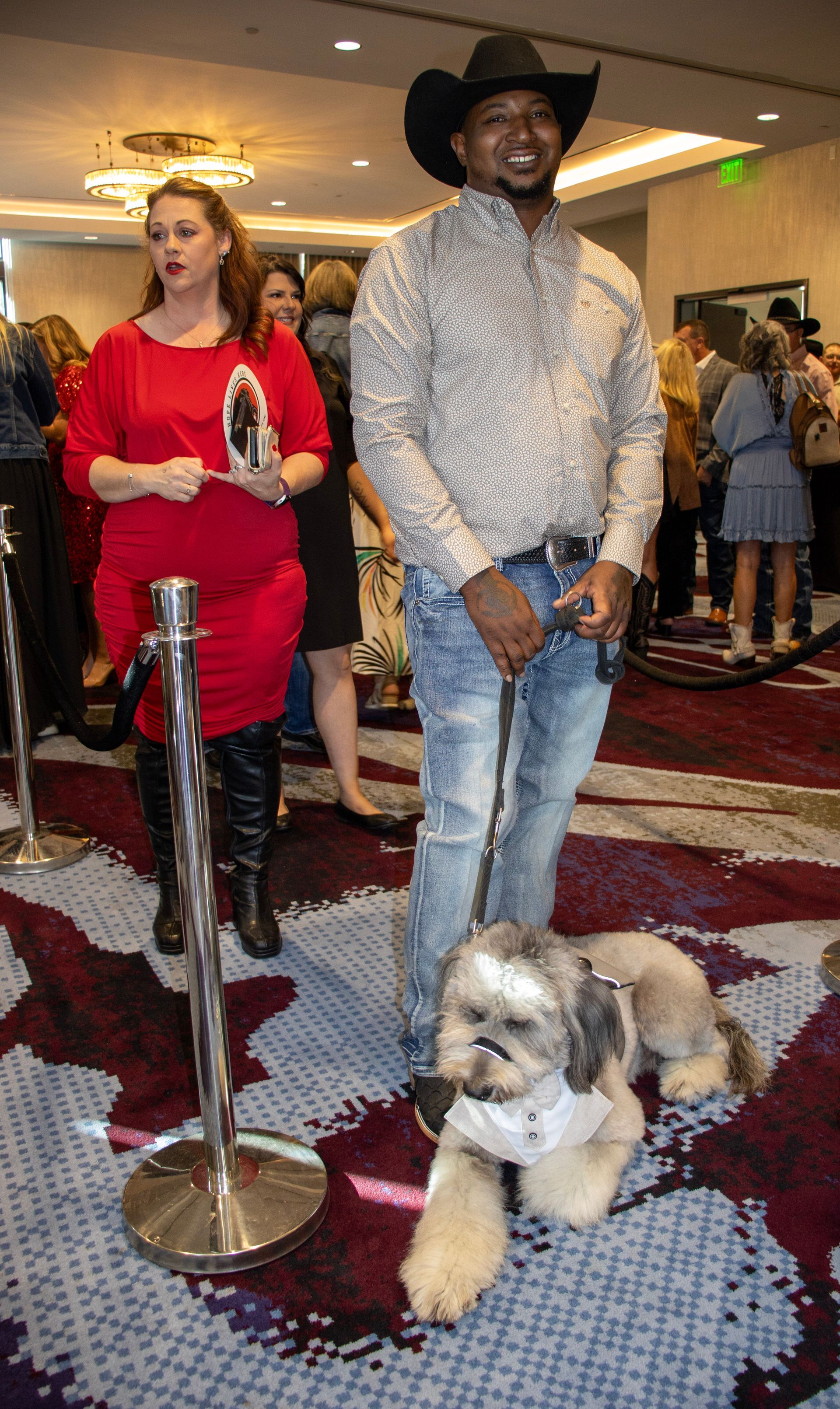 A person in a cowboy hat and patterned shirt stands with a small, leashed dog on a patterned carpet near a stanchion.