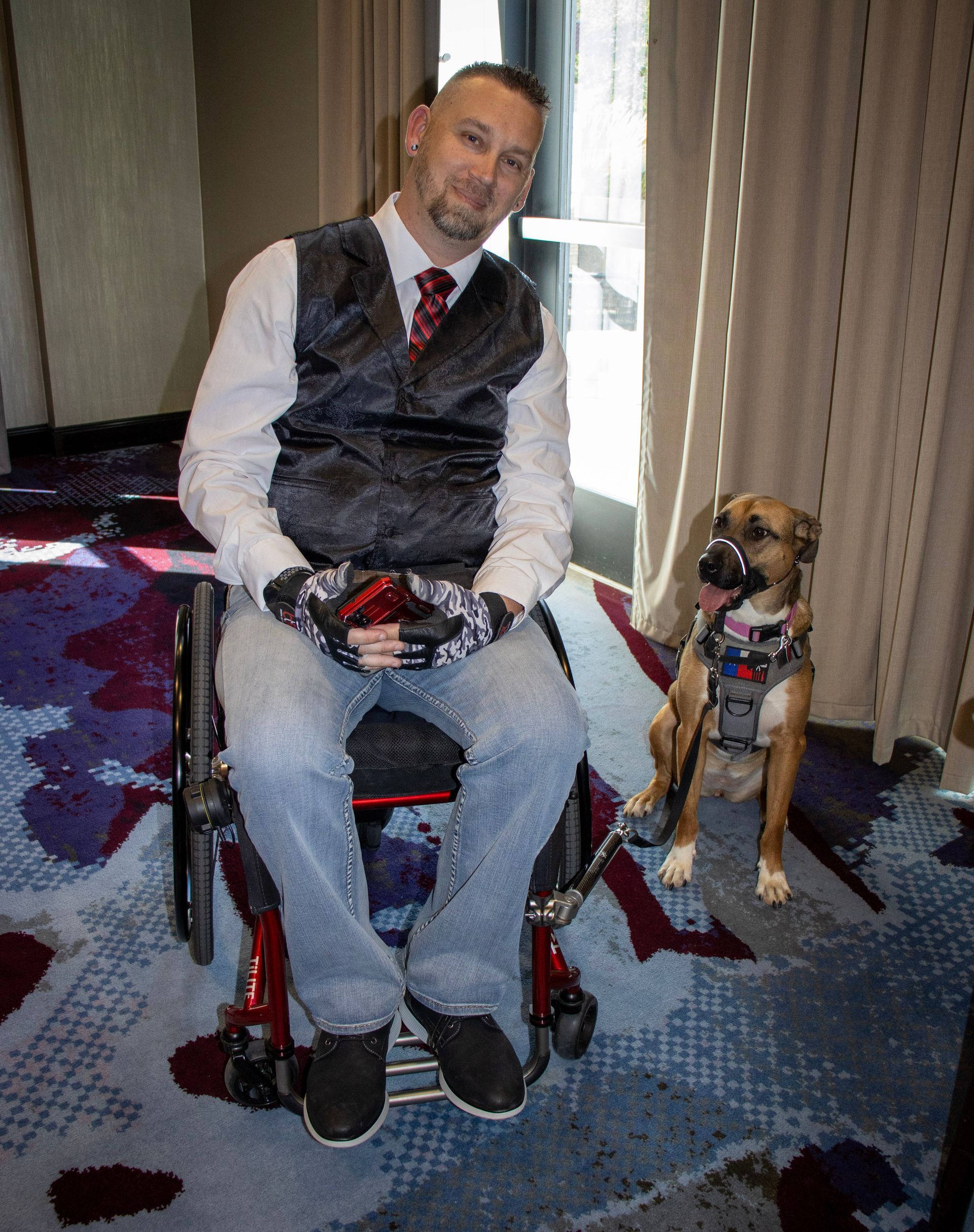 A person in a wheelchair wearing a vest and tie sits next to a service dog wearing a harness near a window with curtains.