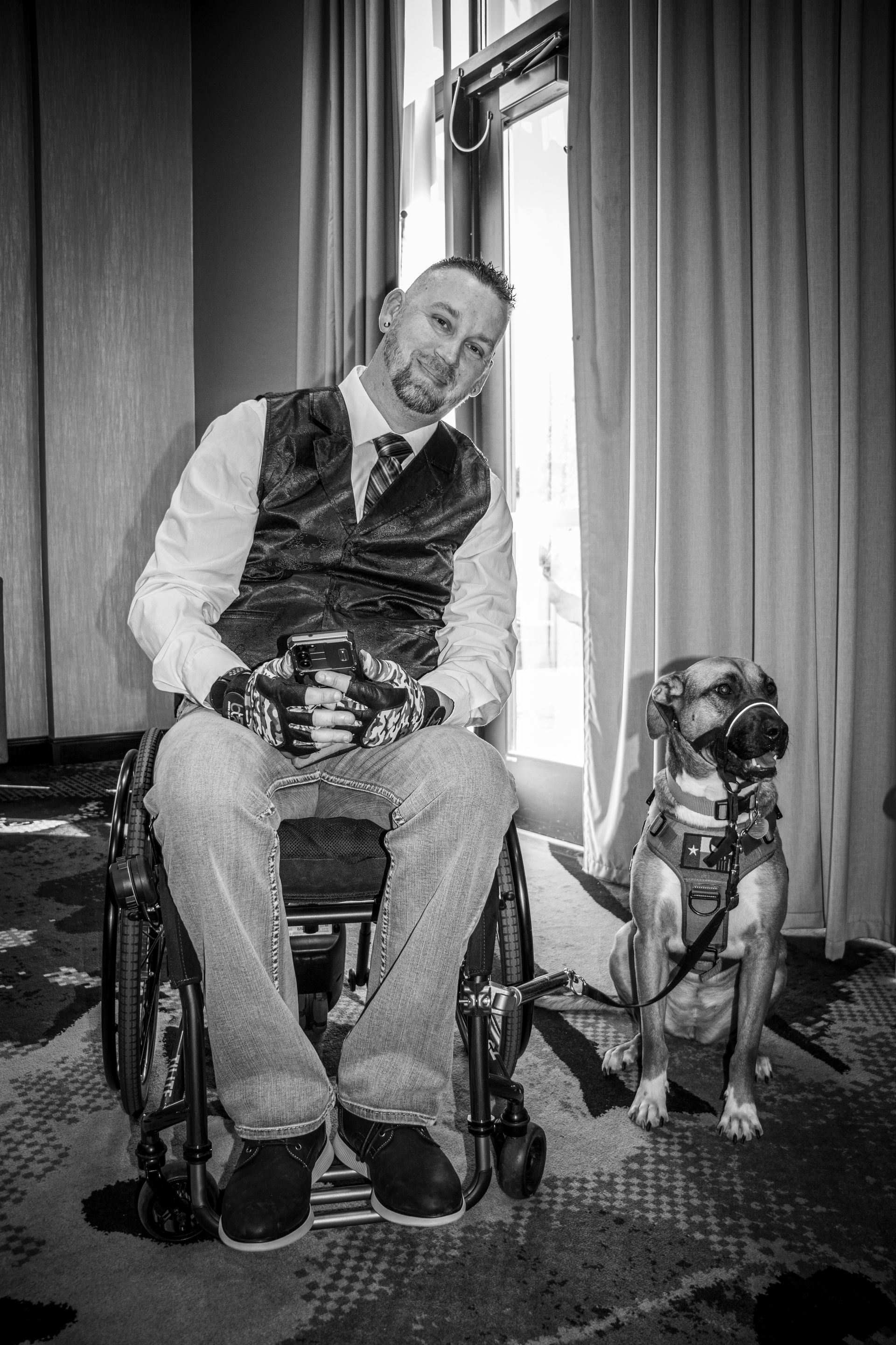 A man in a wheelchair and a service dog sit side by side in front of a window with curtains.