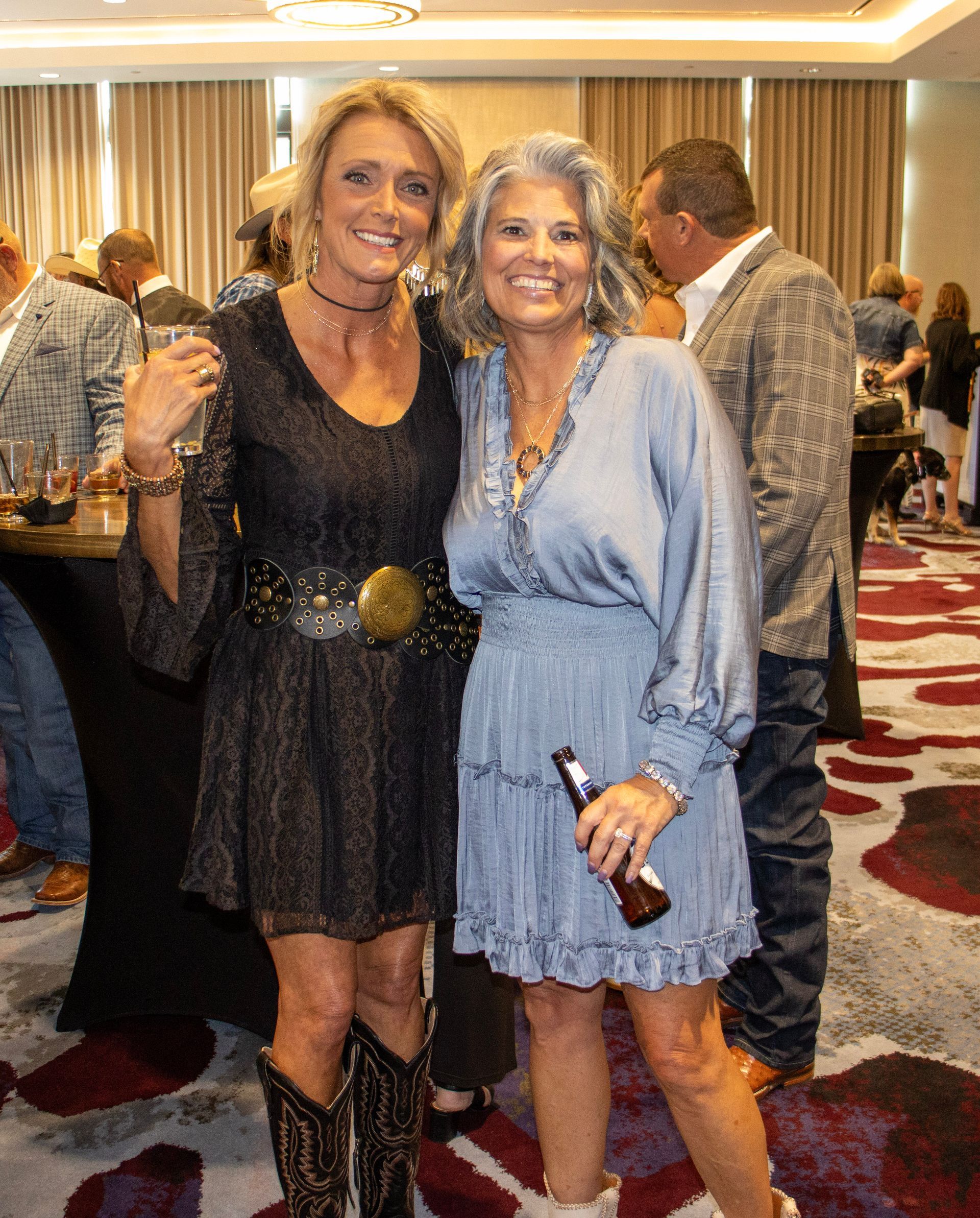 Two smiling people posing at an indoor event, one in a black lace dress and boots, the other in a blue dress holding a beer.