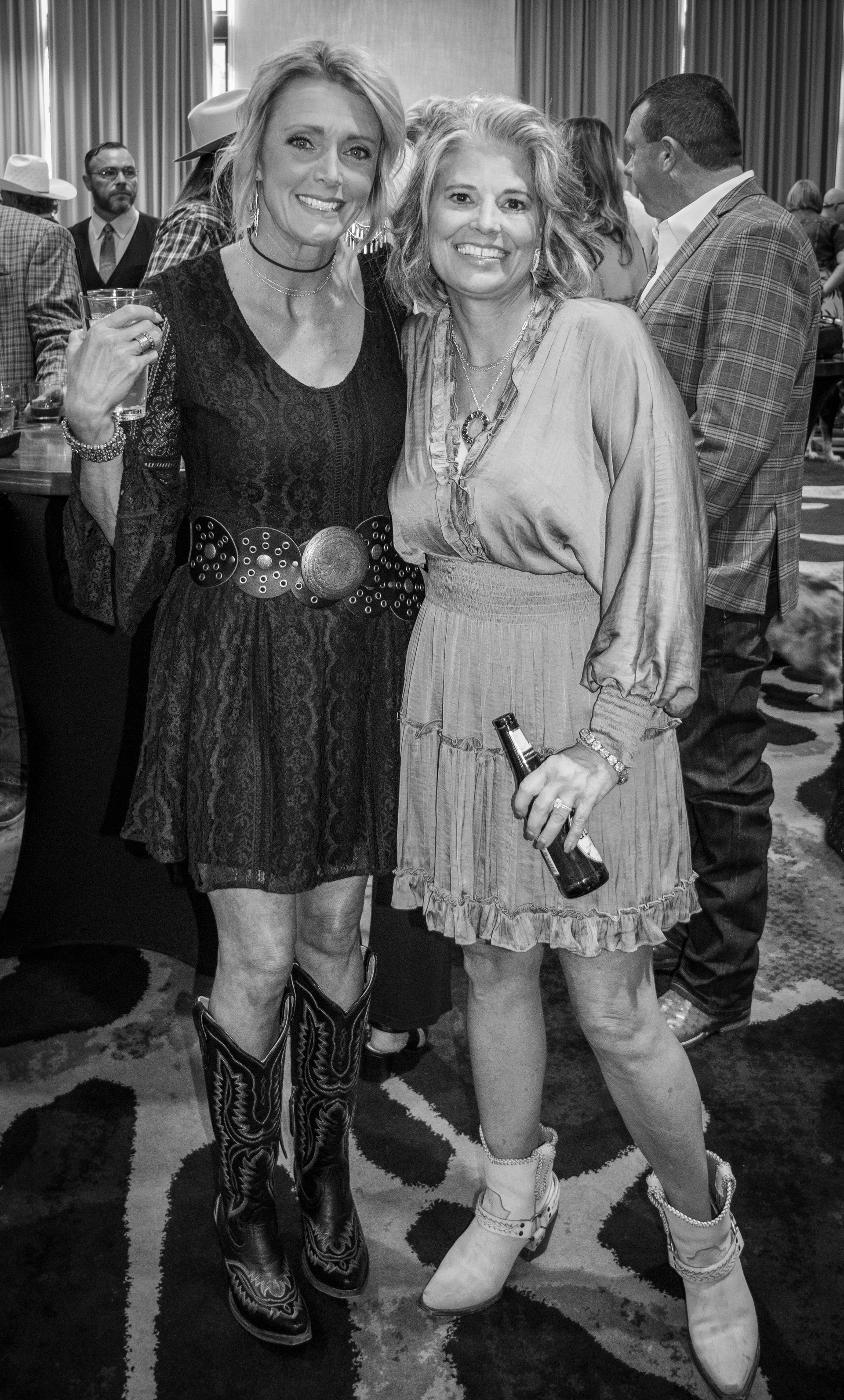 Two smiling women in dresses and cowboy boots stand together at an indoor event, one holding a drink.