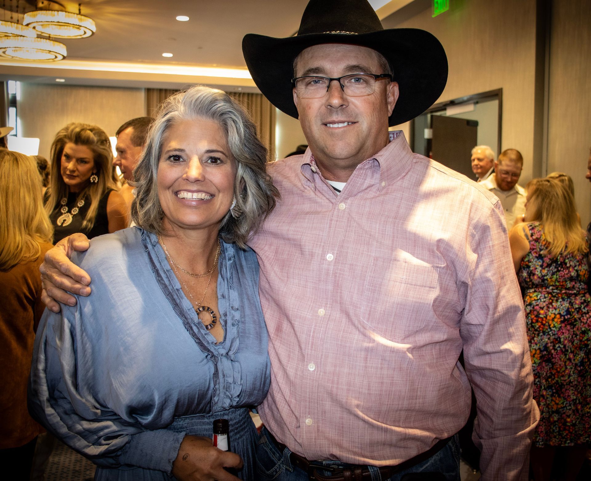 A smiling woman in a blue blouse and a man in a light-colored button-down shirt and cowboy hat stand together at an event.