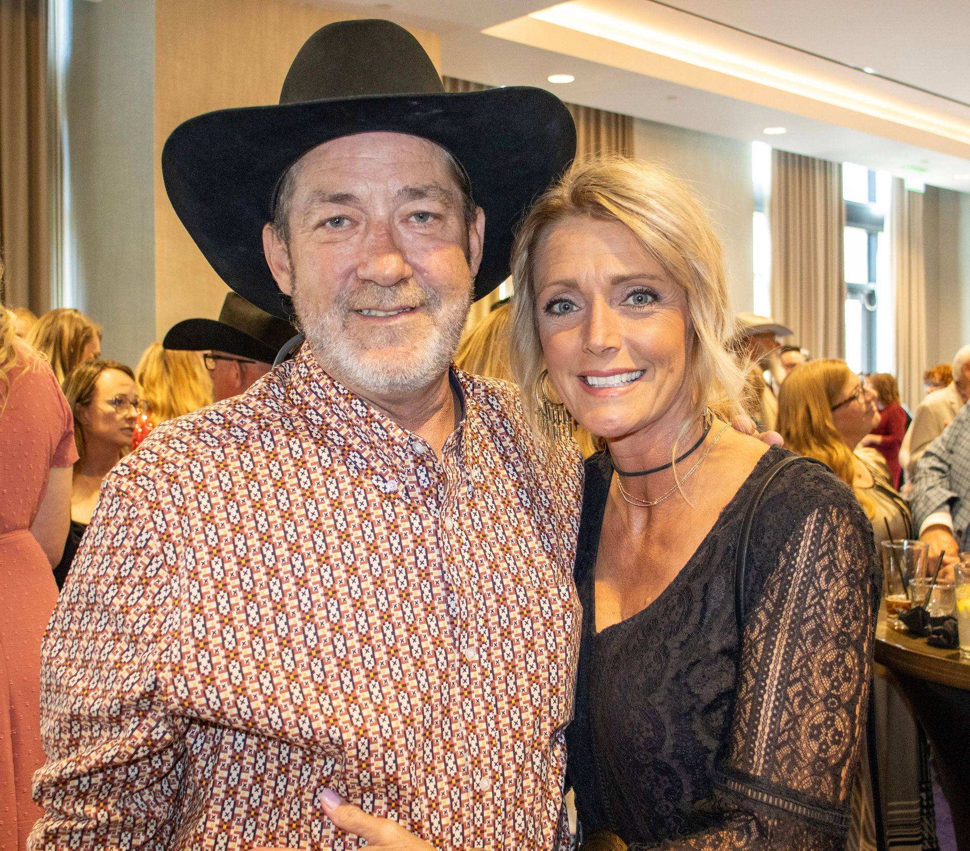 A smiling man in a cowboy hat and patterned button-down shirt stands next to a smiling woman in a black lace top.