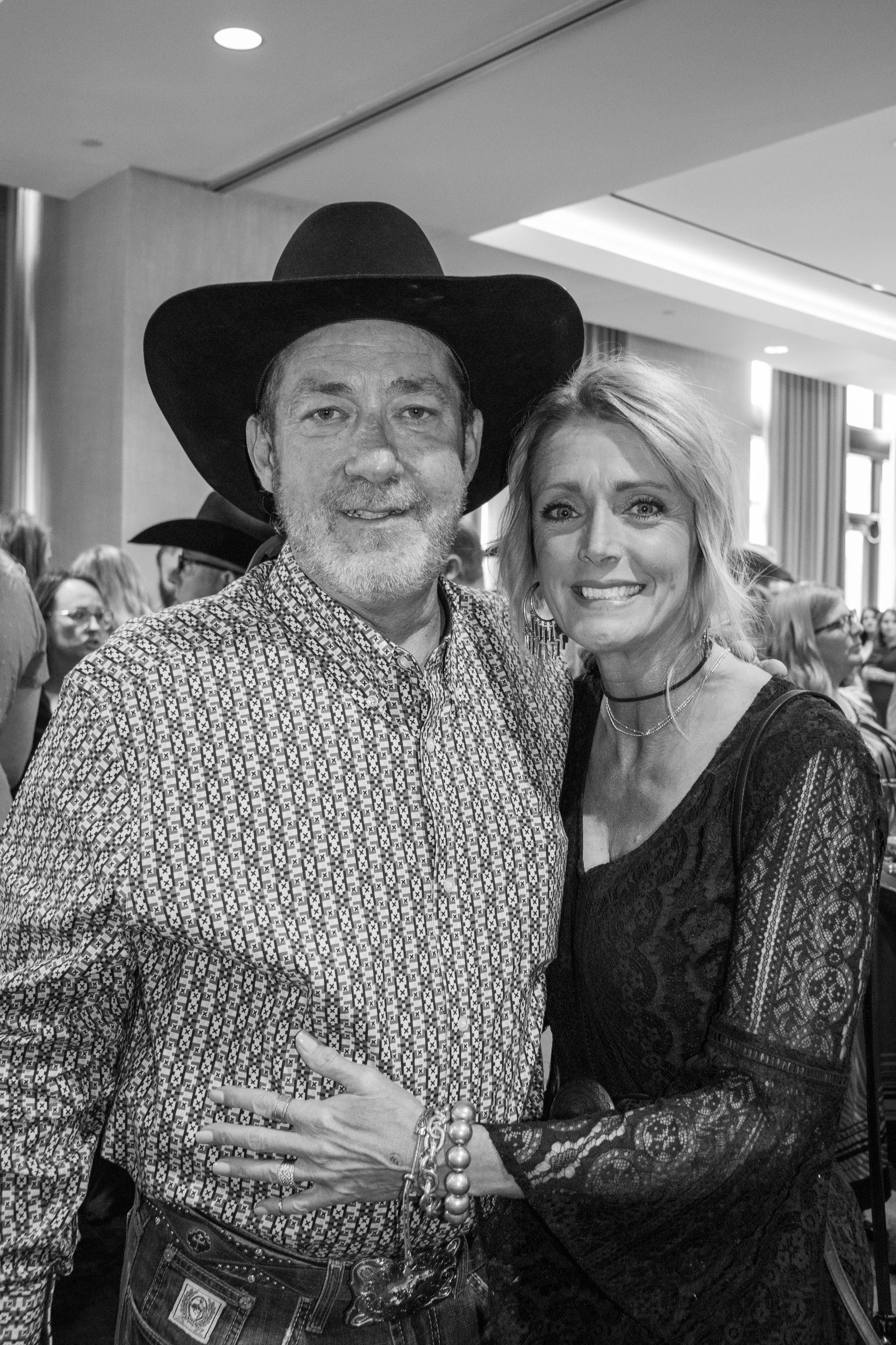 A man in a cowboy hat and patterned shirt poses with a smiling woman in a dark lace top at an indoor event.