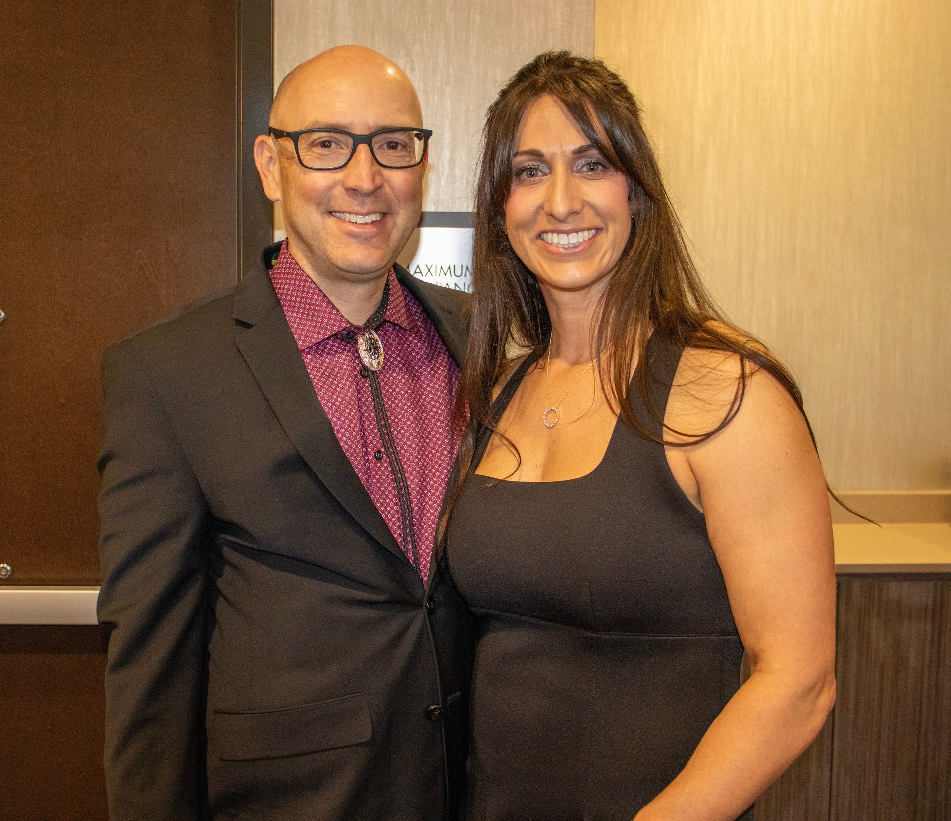 A man in a suit and bolo tie stands next to a woman in a black dress, both smiling at the camera.