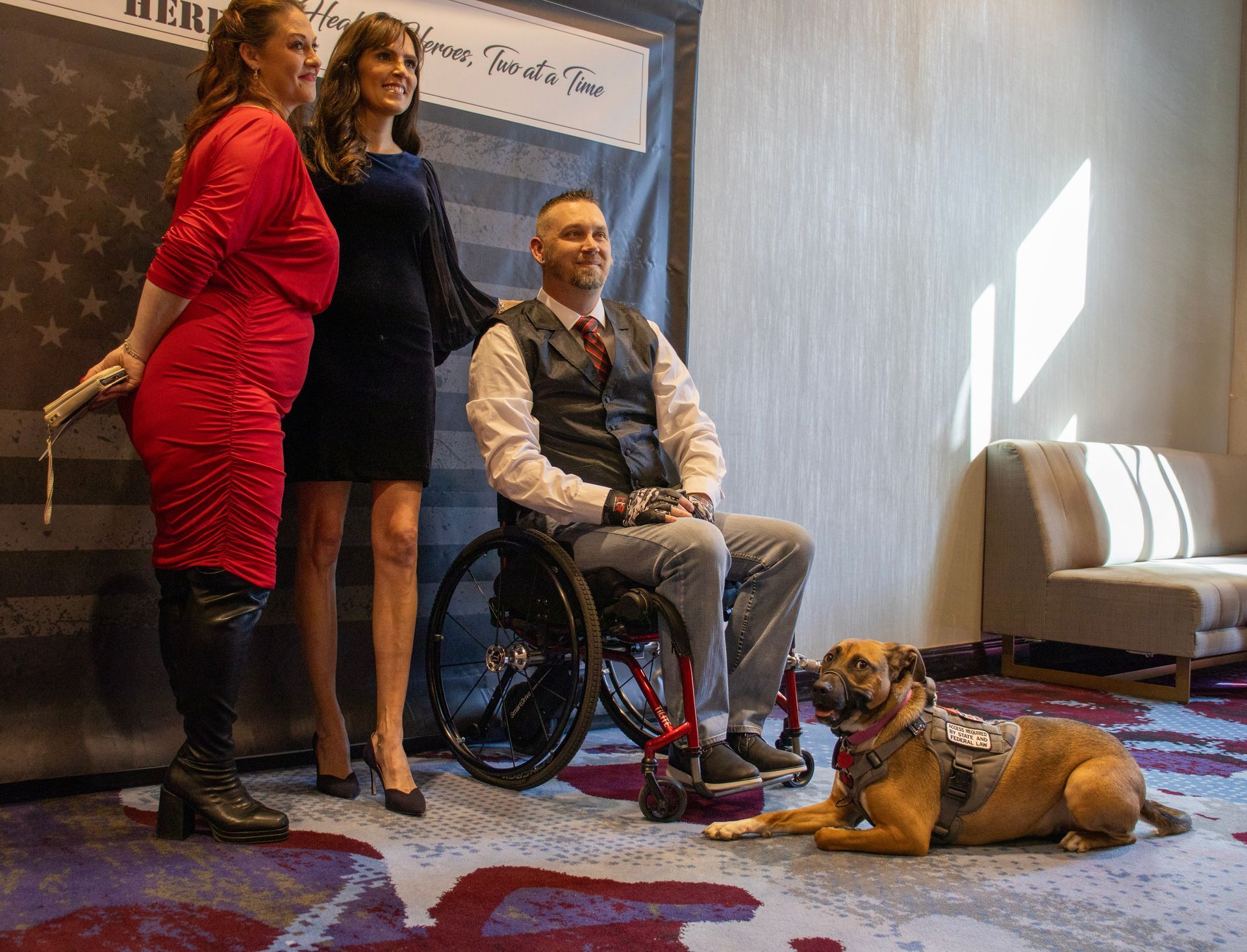 Two women and a man in a wheelchair with a service dog pose in front of a flag-patterned backdrop.