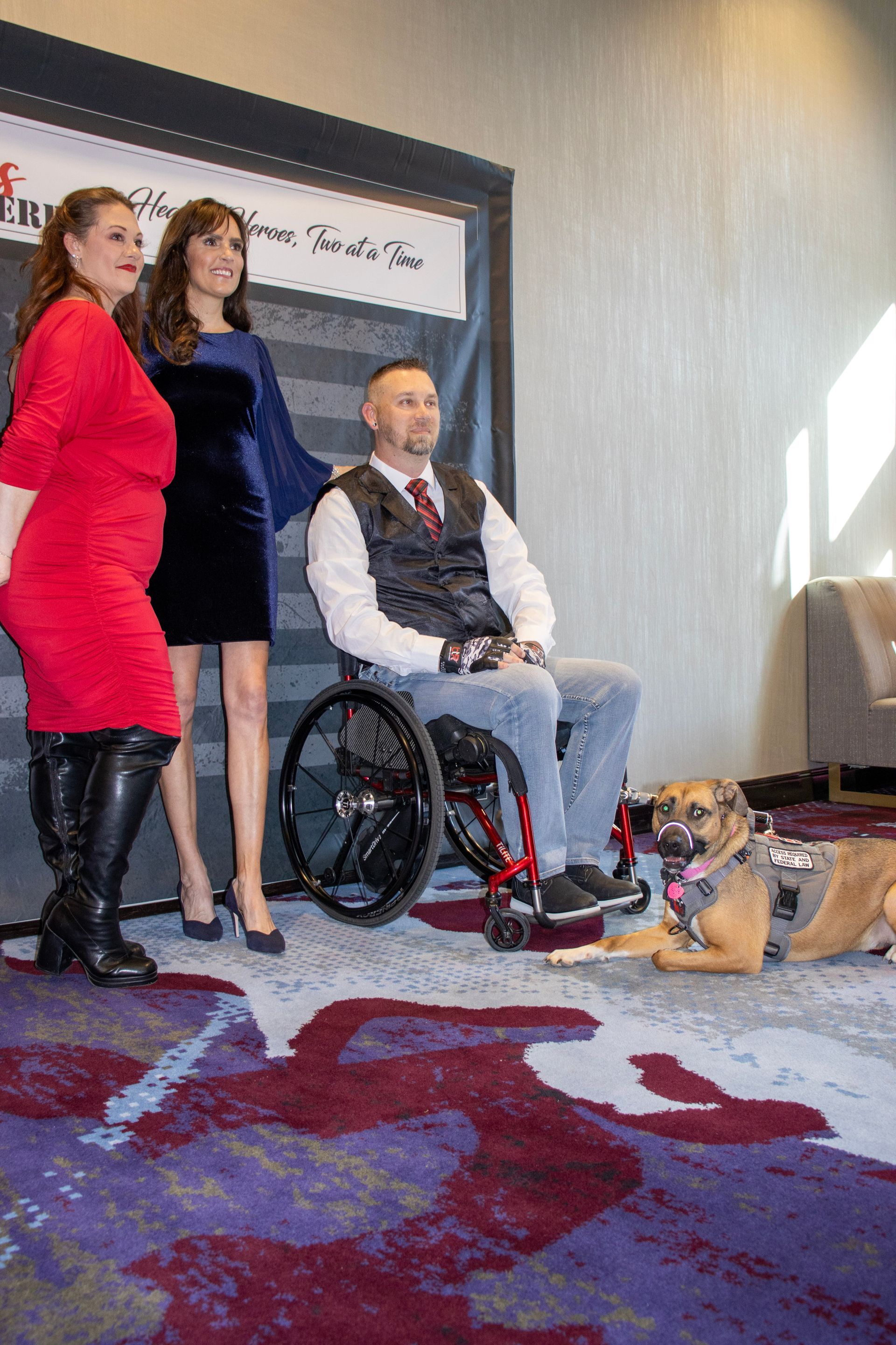 Two women and a man in a wheelchair posing with a service dog in front of a branded event backdrop.