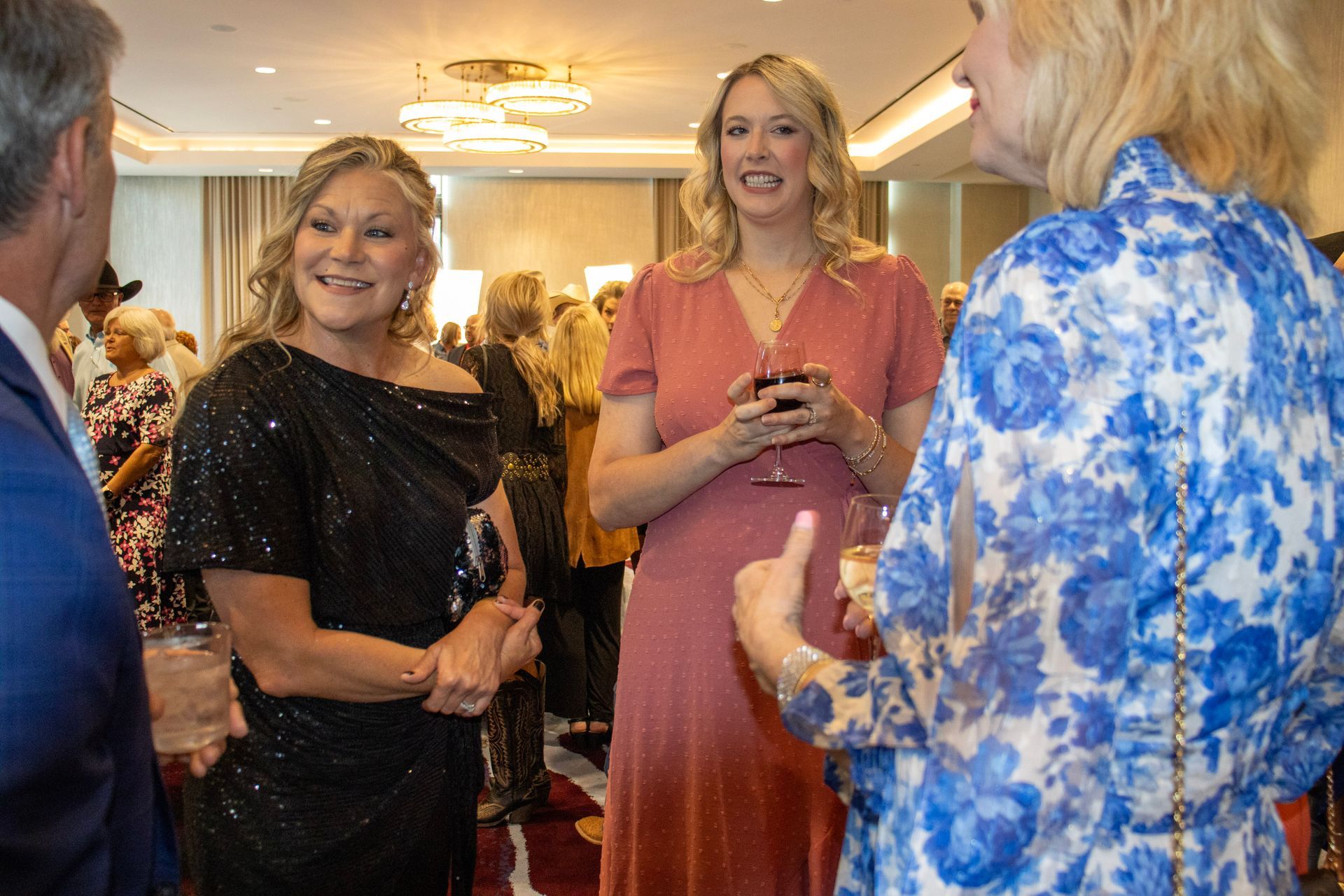 Three people talk at an indoor gathering; one wears a black glittery top, another a dusty pink dress, and one a blue print.