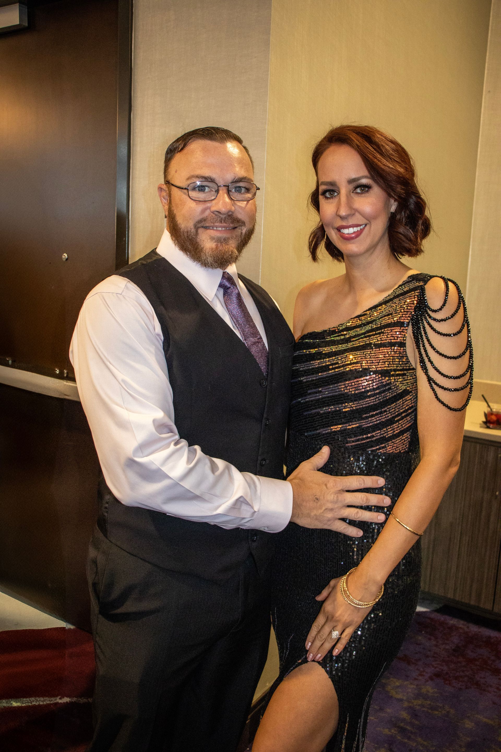 A smiling man in a vest and tie stands with an arm around a smiling woman in a one-shoulder, sequined black dress.