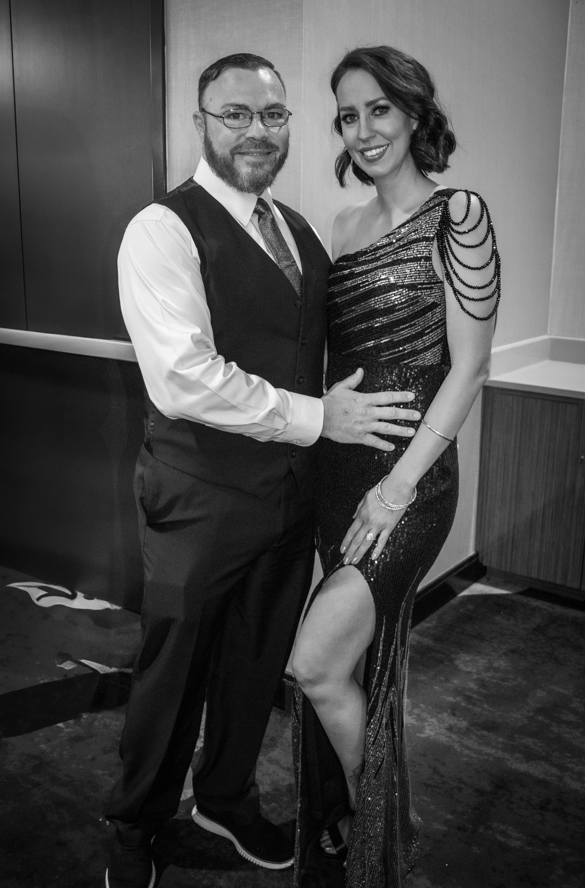 A man in a vest and a woman in a sequined, one-shouldered gown posing together for a formal event.