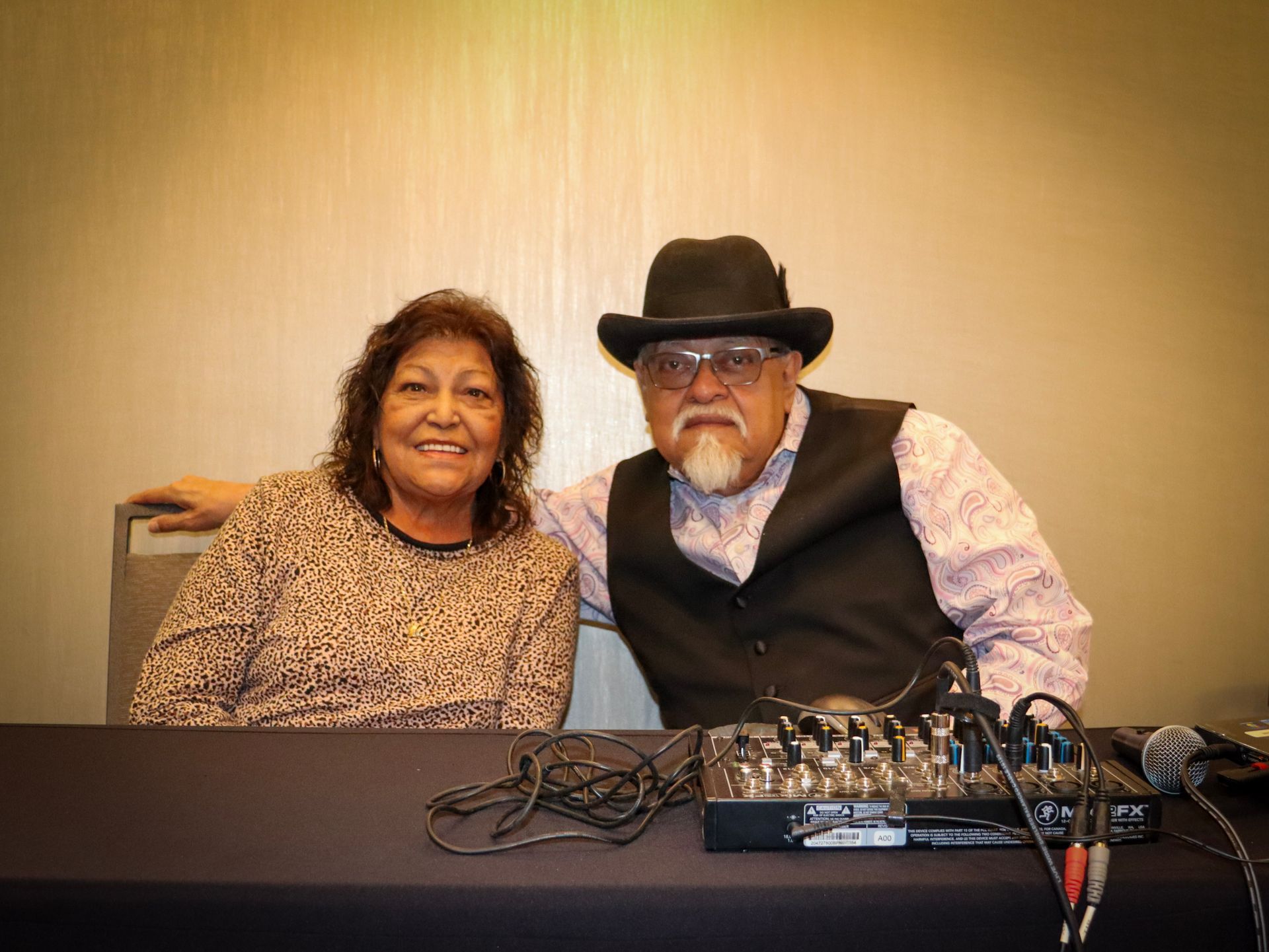 A smiling couple sits side-by-side at a table with an audio mixer, against a plain wall.
