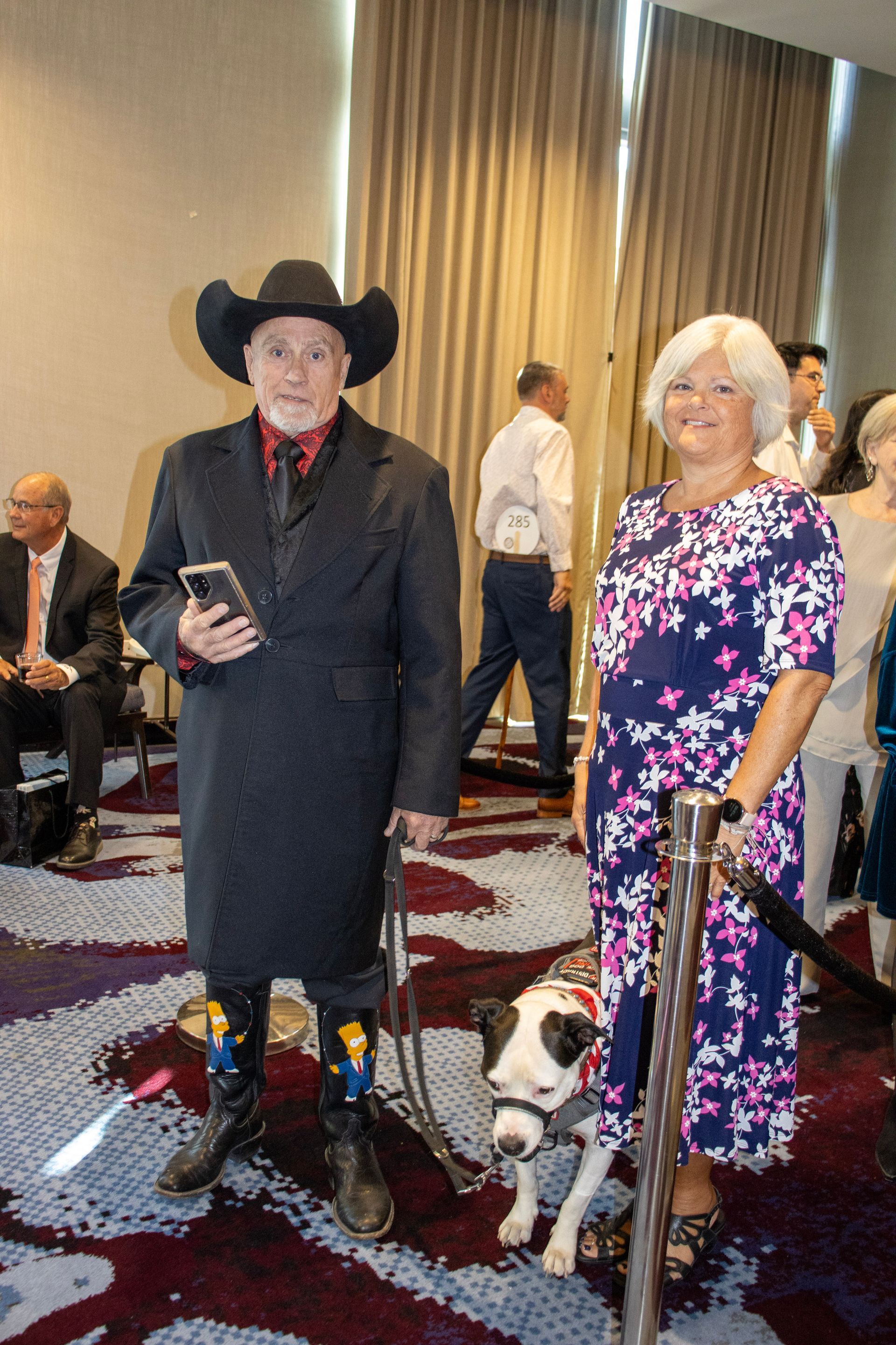 A person in a black coat and cowboy hat stands next to someone in a floral dress and a service dog in a carpeted room.