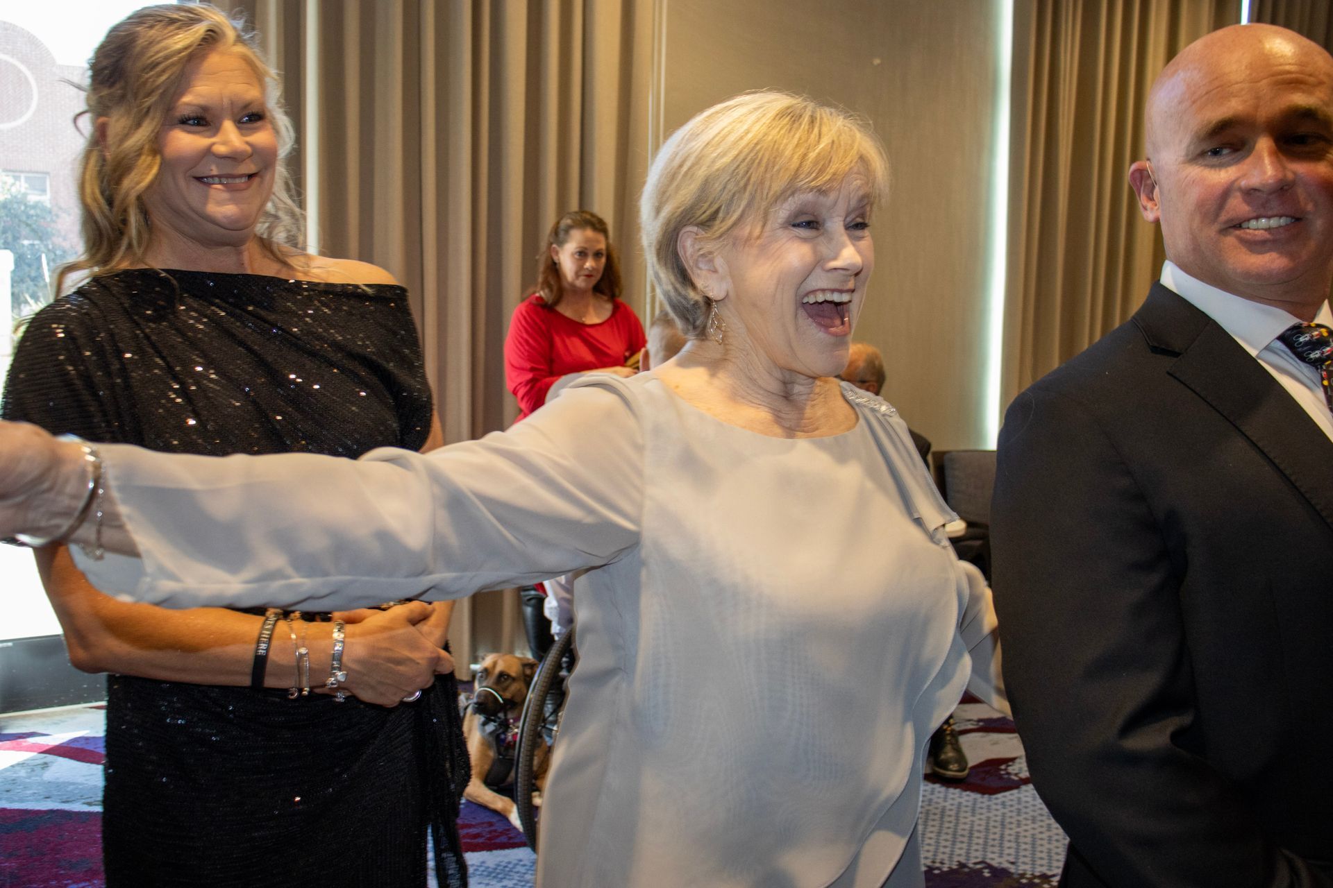 Three people smile indoors, with a person in a grey top gesturing forward in the foreground.