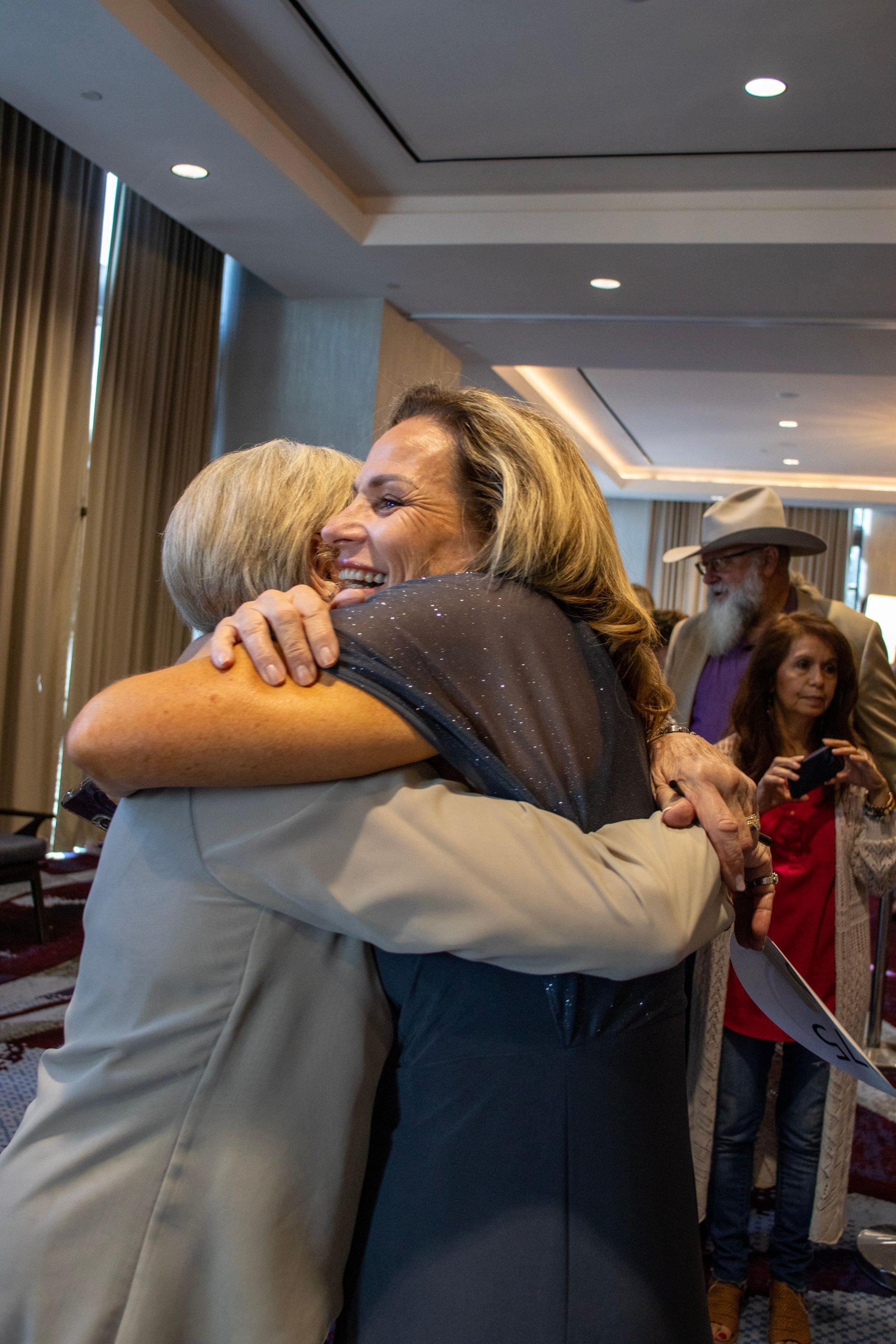 Two people embrace warmly in a conference room while others stand in the background.