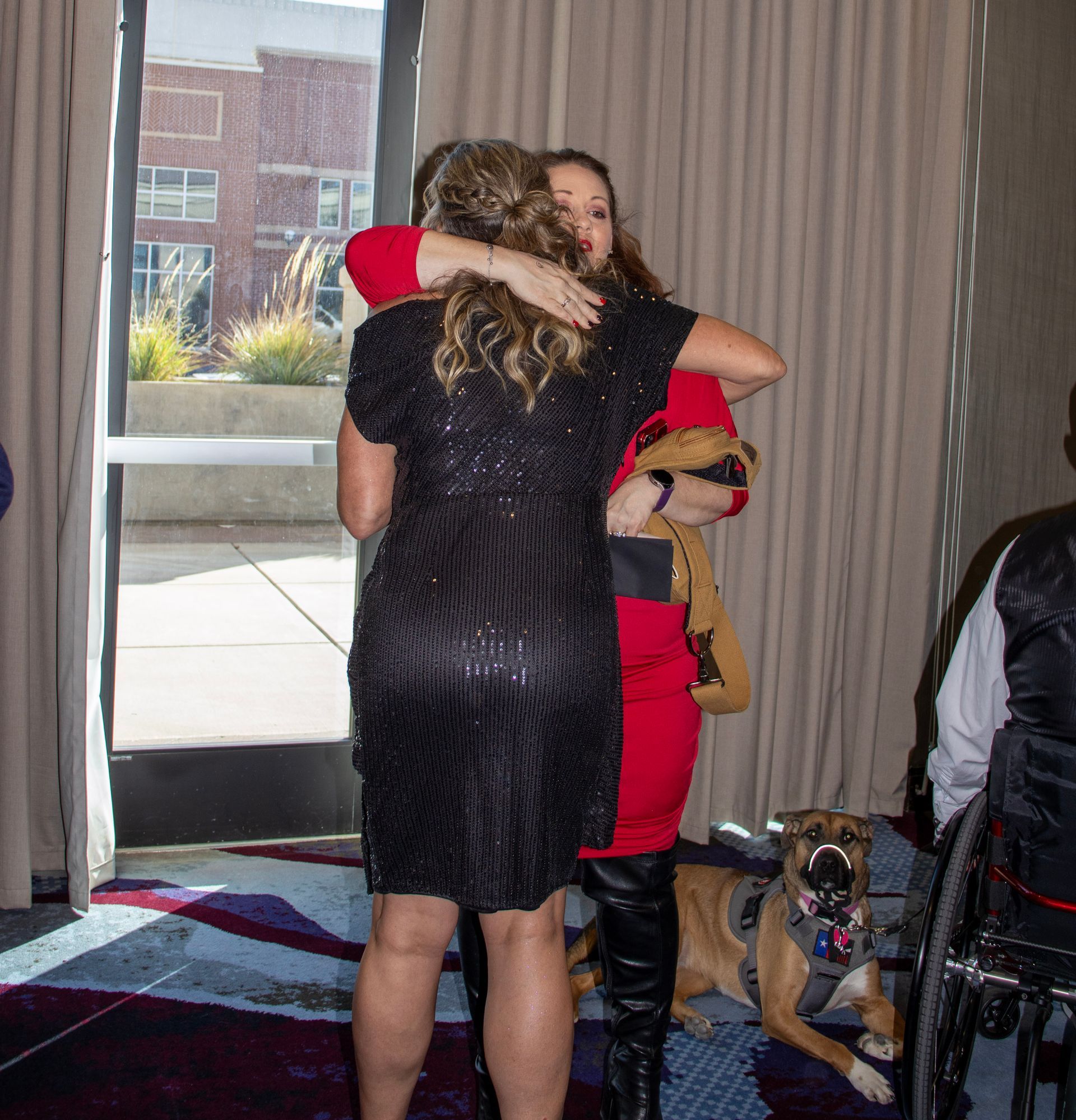 Two people embrace in a room near a window. A dog wearing a harness sits on the floor beside them near a wheelchair.