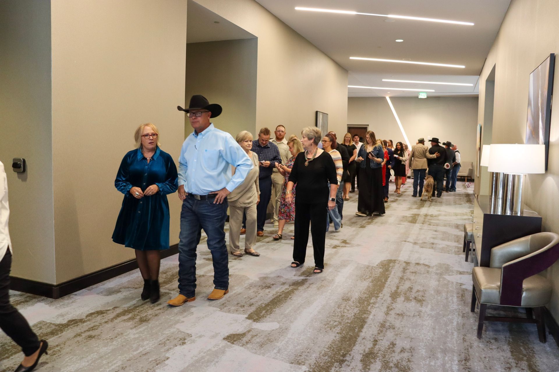 A crowd of people walks down a hallway in a hotel or convention center with patterned carpet and modern lighting.