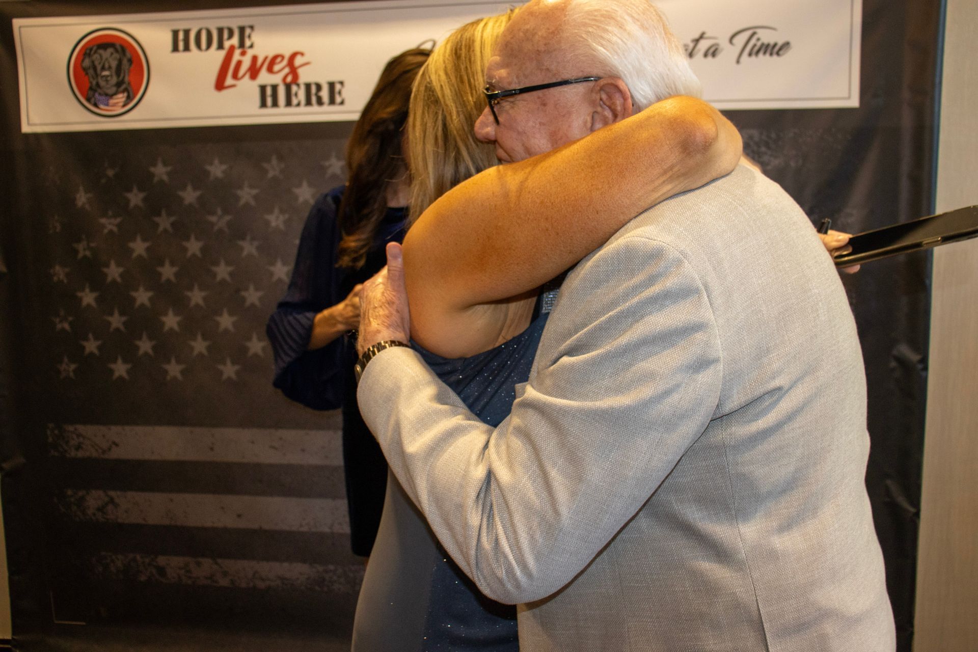 Two people share a warm embrace in front of a banner that reads 
