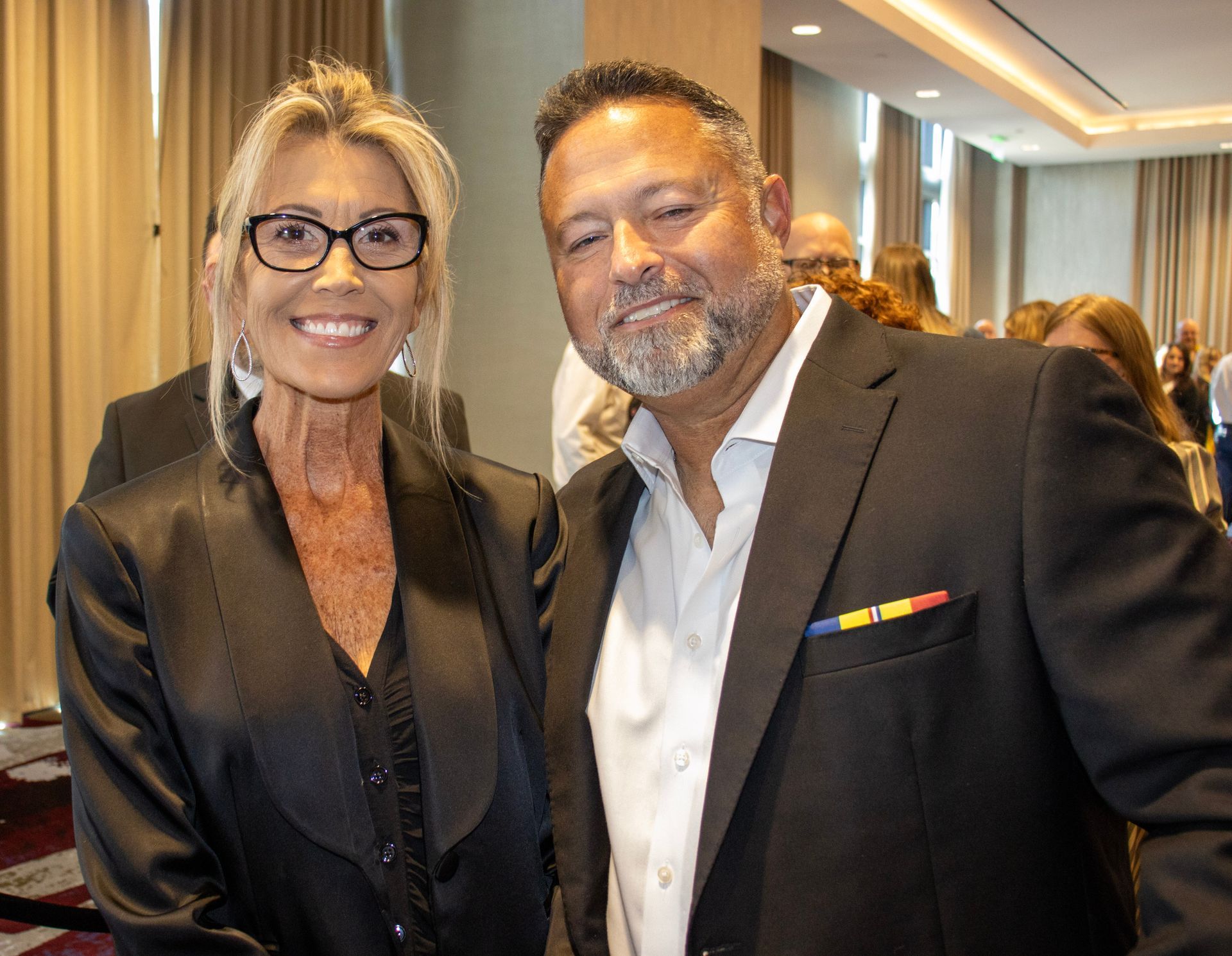A woman wearing glasses and a dark blazer smiles next to a man with a beard in a dark suit with a colorful lapel pin.