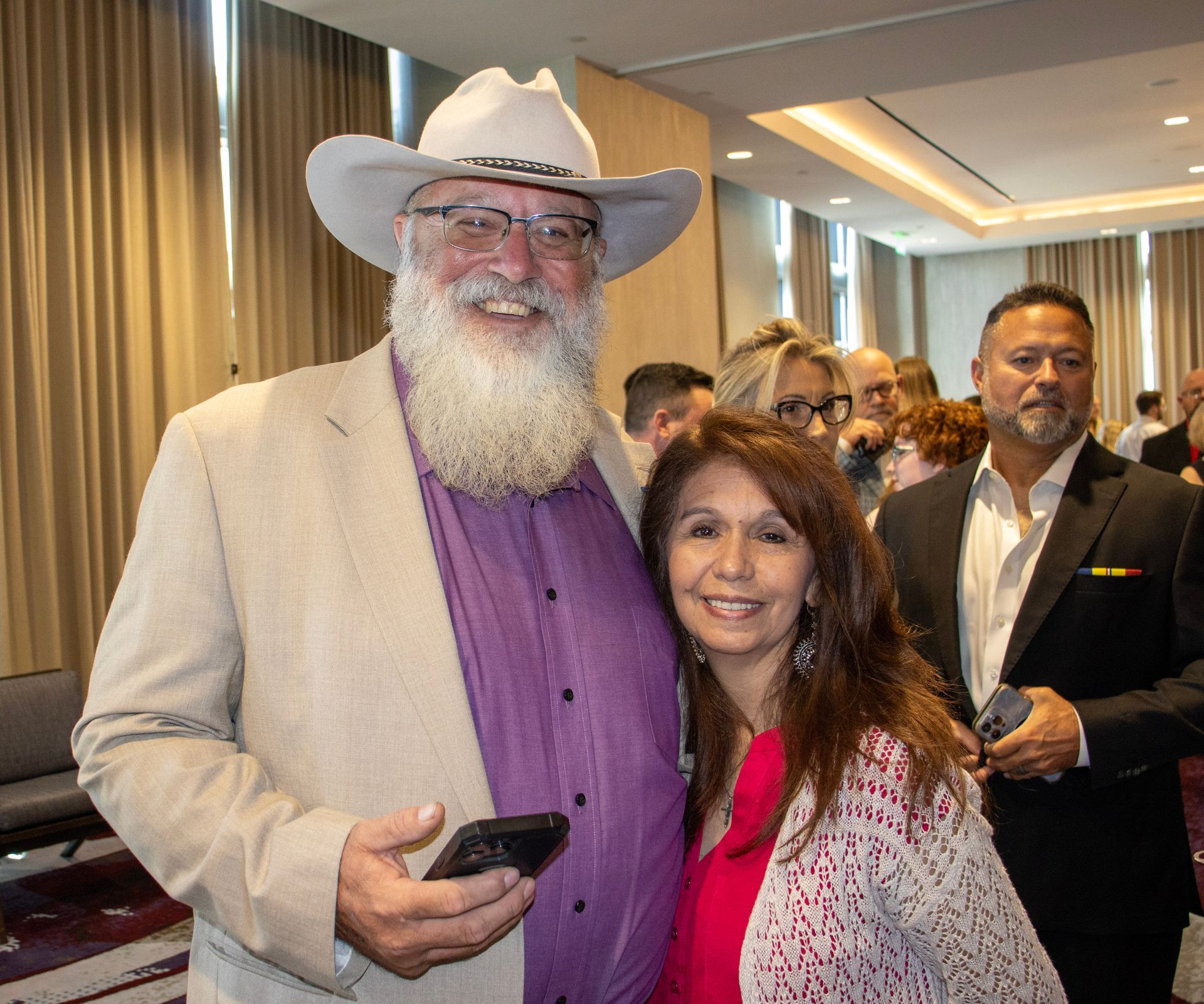 A man in a cowboy hat and a woman in a pink top smile for a photo at an indoor event, with a man standing nearby.