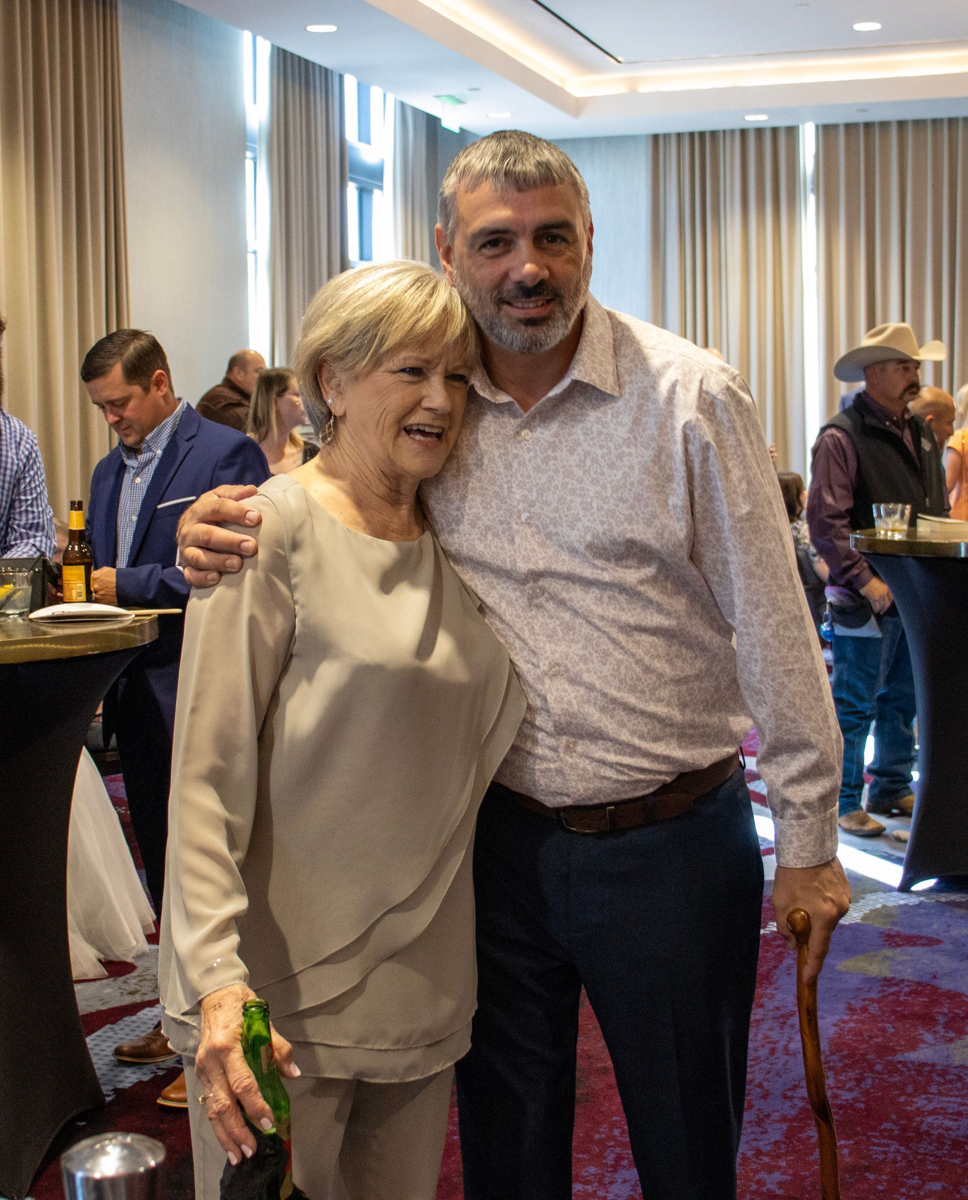 A smiling woman in a beige top and a man in a patterned shirt embrace in a crowded, indoor event venue.