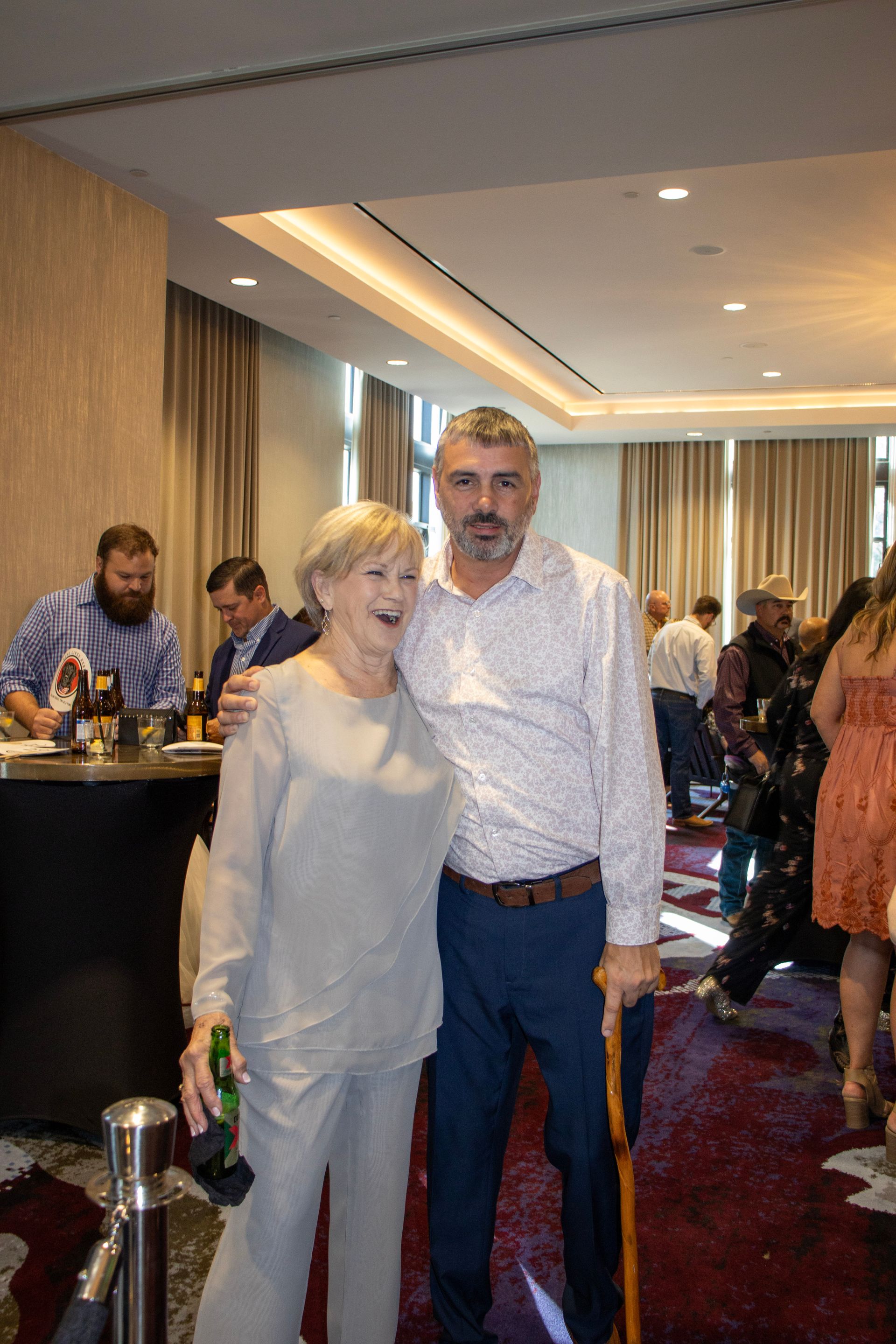 A man and woman smiling in a conference room, with guests blurred in the background. The man holds a walking cane.