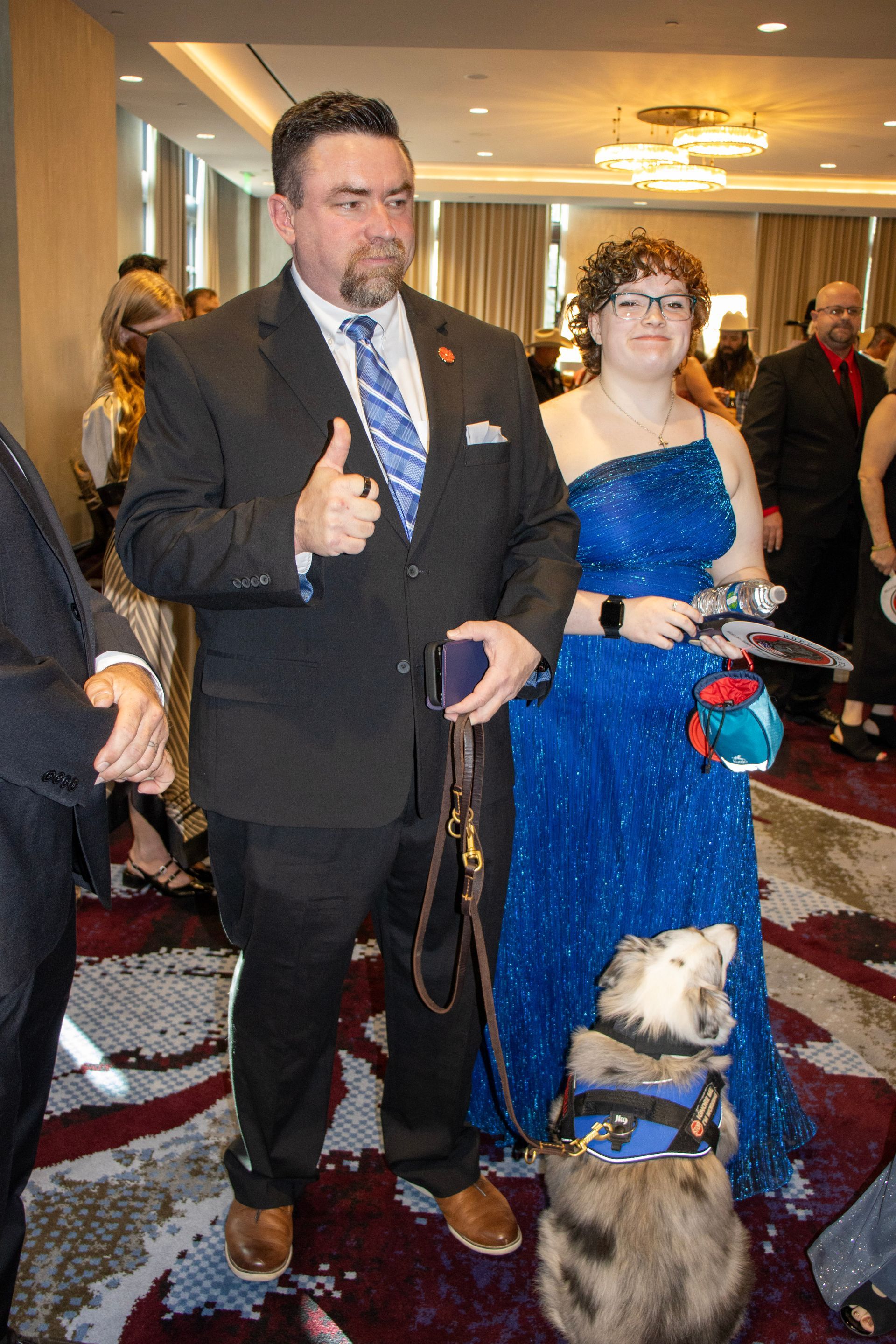 A person in a suit giving a thumbs up and a person in a blue gown stand with a service dog at an indoor event.