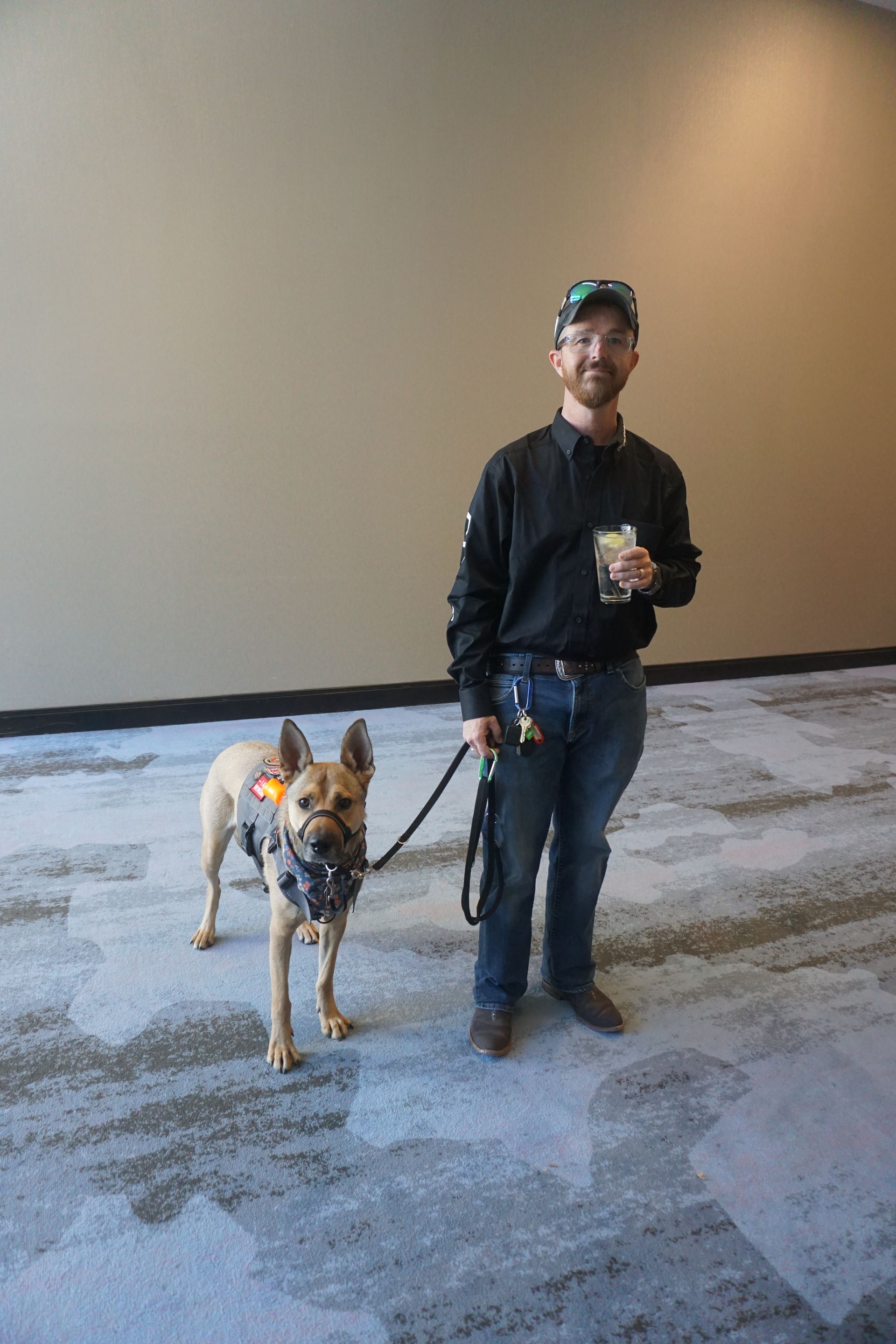 A person in a black shirt and jeans holds a leash attached to a service dog in a vest, standing on a patterned carpet.
