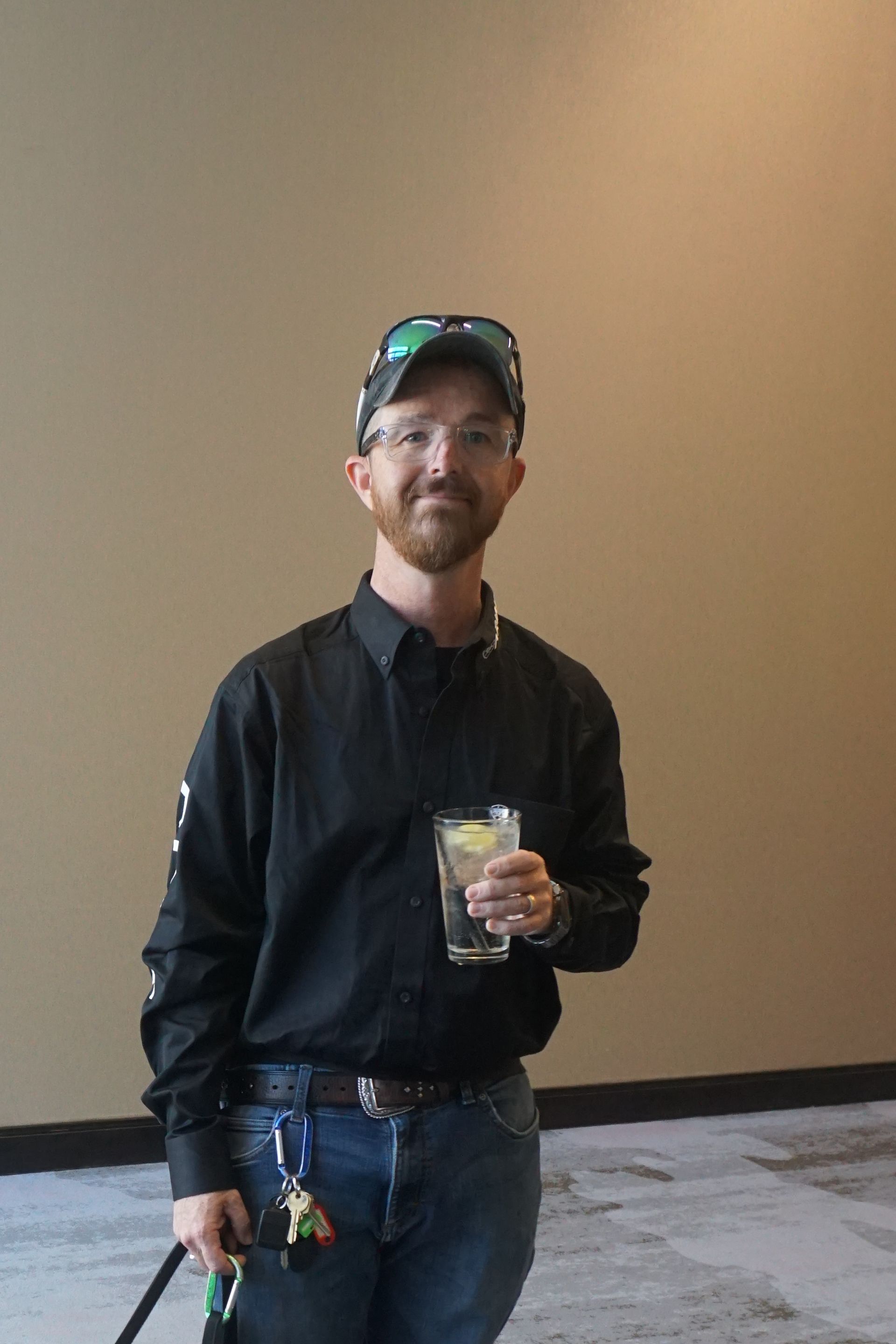 A person wearing a black button-down shirt, jeans, and a cap holds a glass of water while standing against a tan wall.