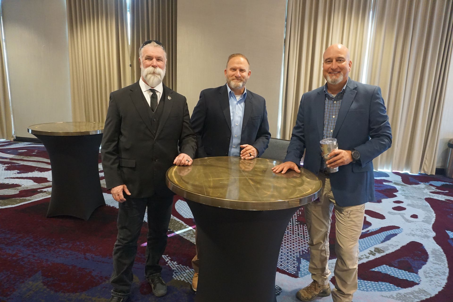 Three men in business attire stand smiling around a tall, circular, marble-topped table in a hotel conference room.