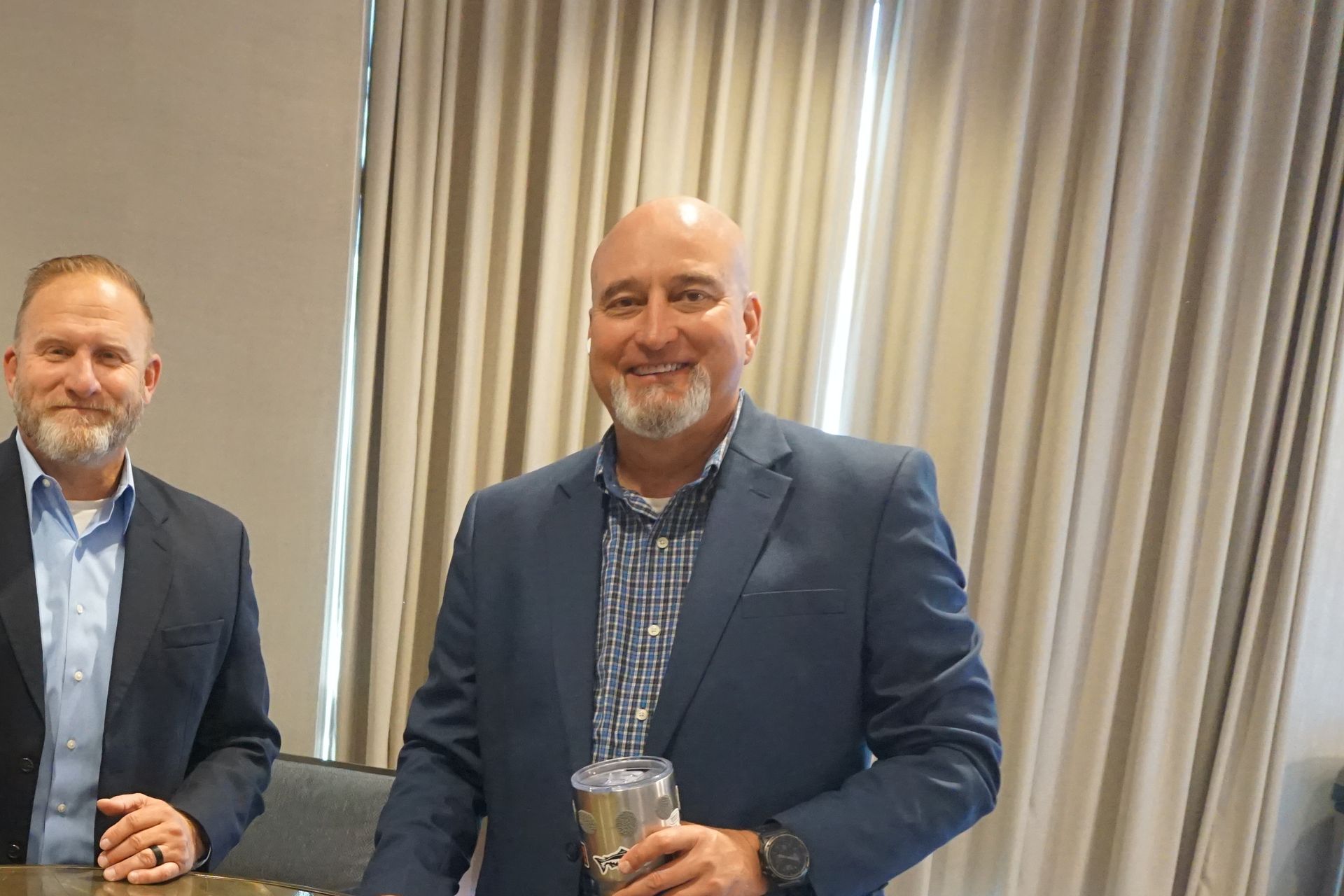 Two people with beards, dressed in blazers and button-down shirts, stand smiling in front of vertical curtains.