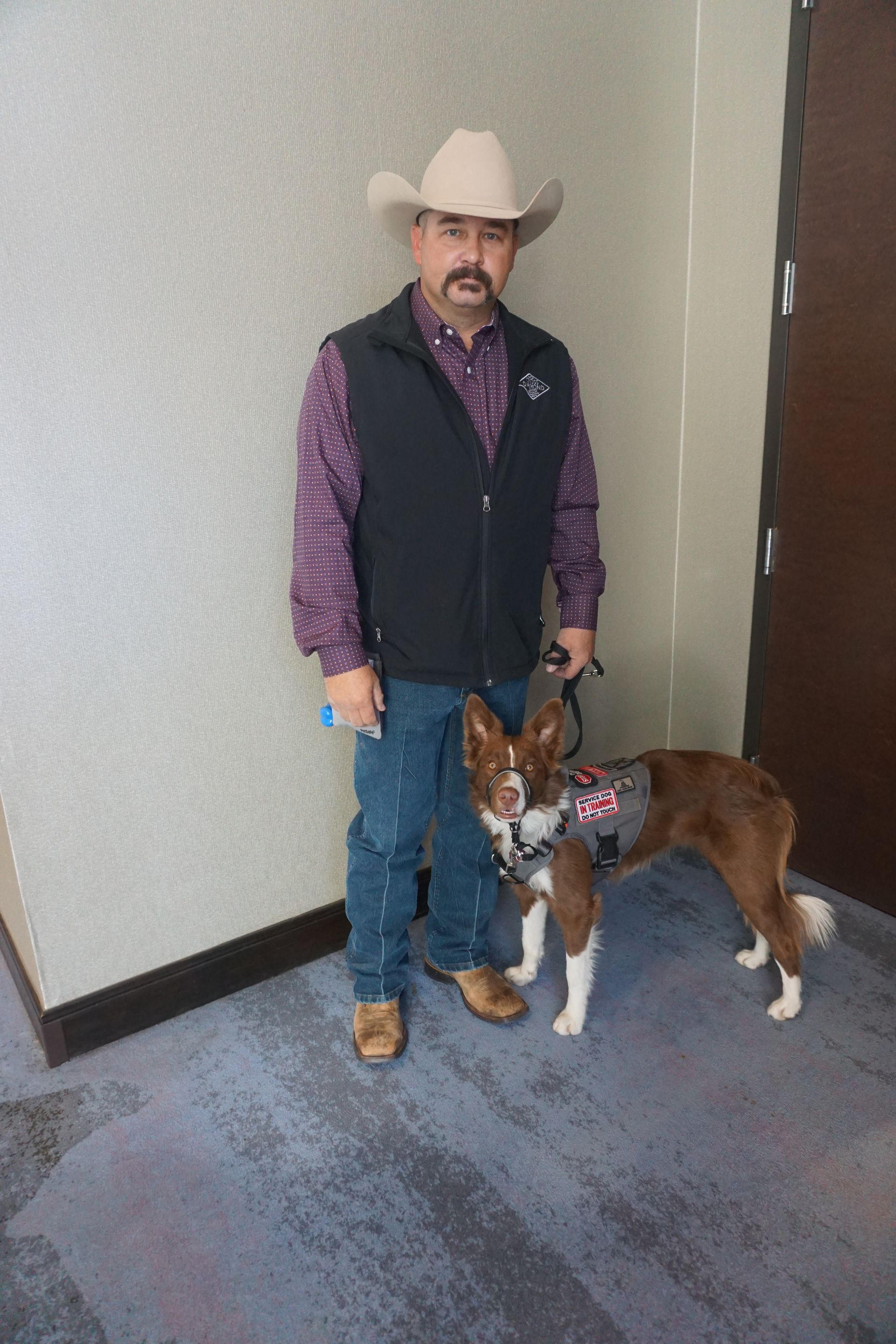 A man in a cowboy hat and vest stands indoors with a brown and white dog wearing a service harness.