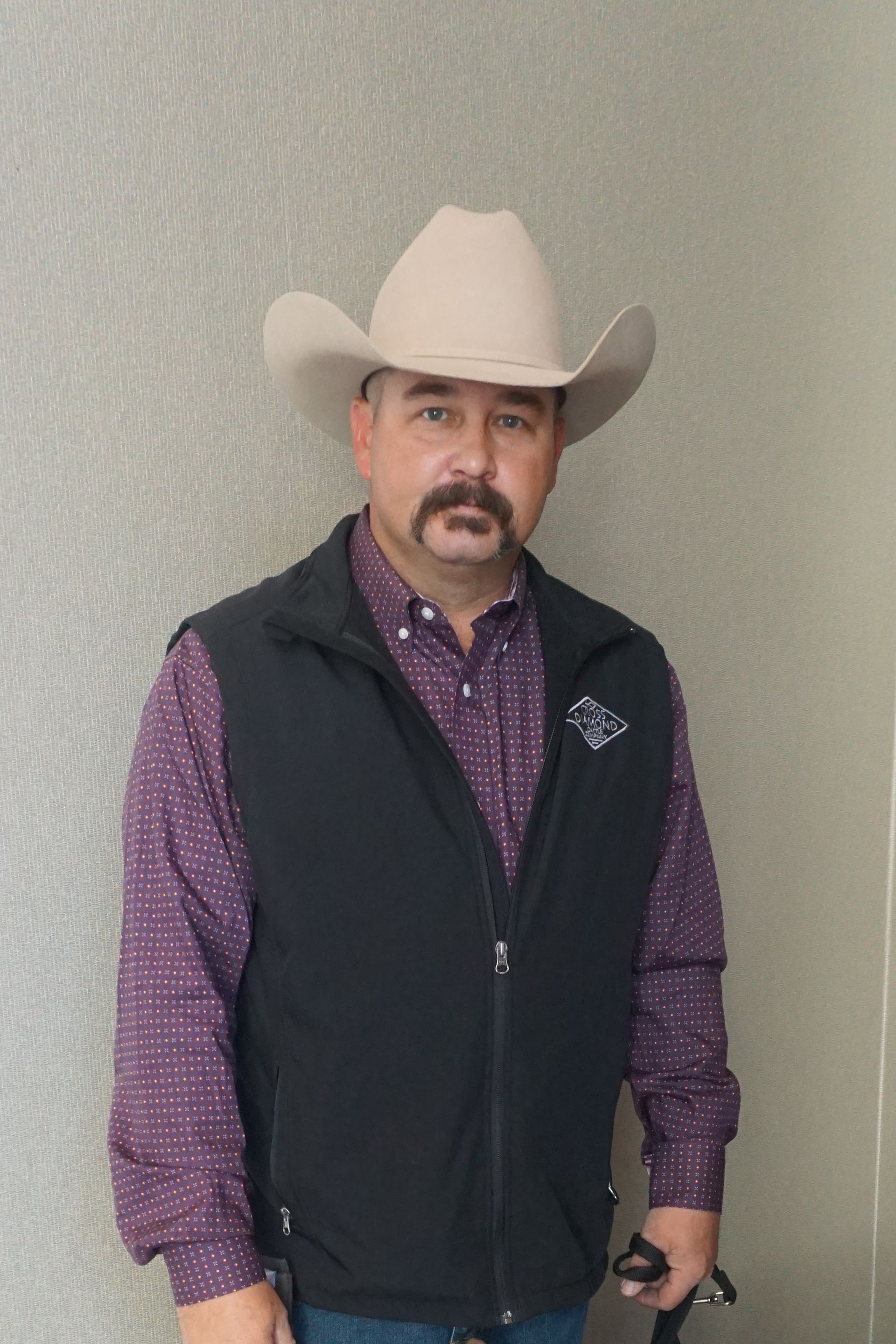 A person wearing a light-colored cowboy hat, a purple patterned shirt, and a black vest stands against a textured wall.