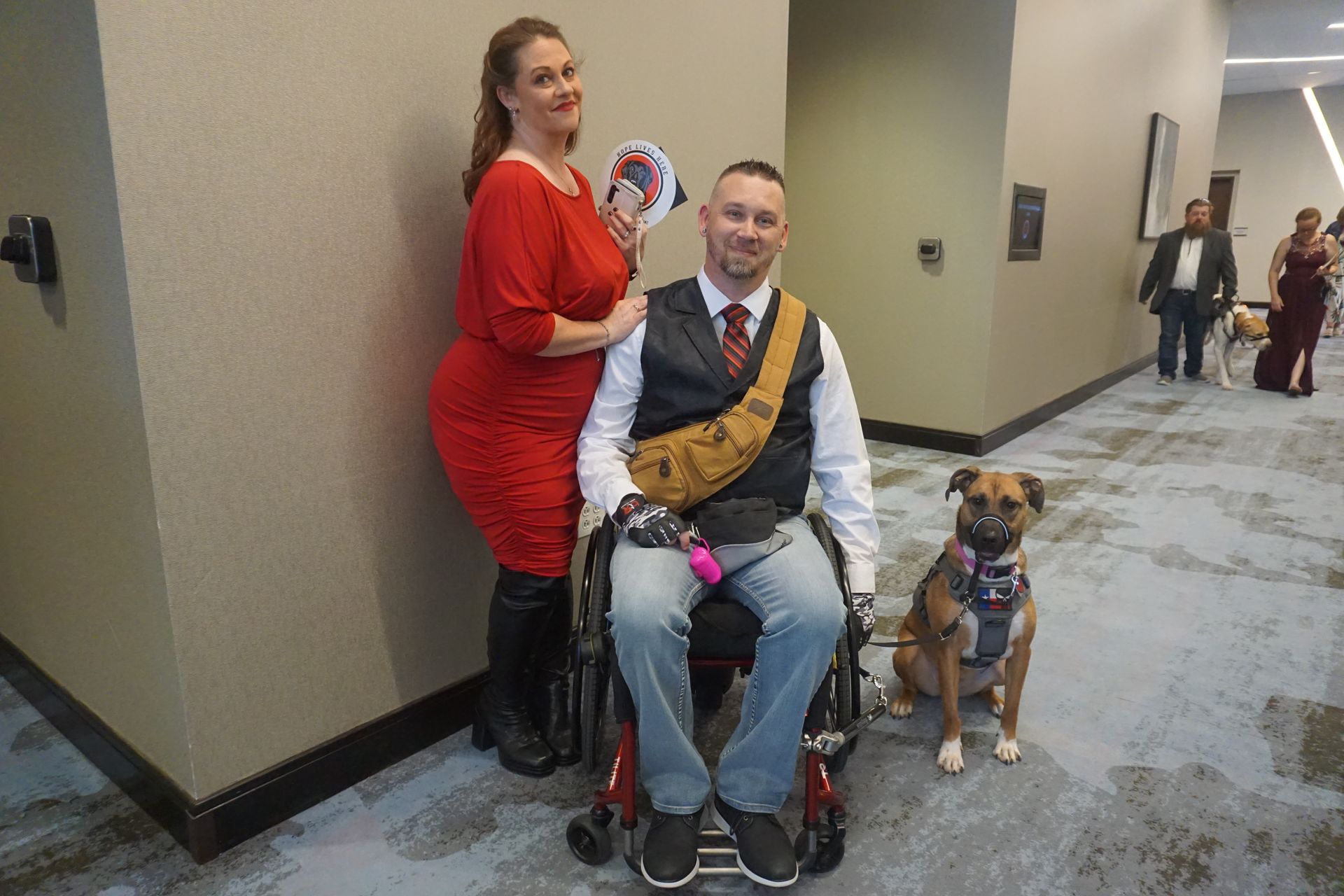 A person in a red dress stands beside a man in a wheelchair and vest, with a dog sitting nearby in a hotel hallway.