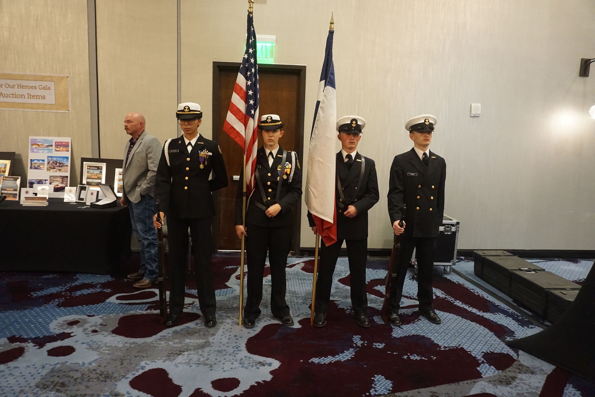 A four-person color guard in navy uniforms stands in a hotel ballroom, holding the U.S. and Texas flags.