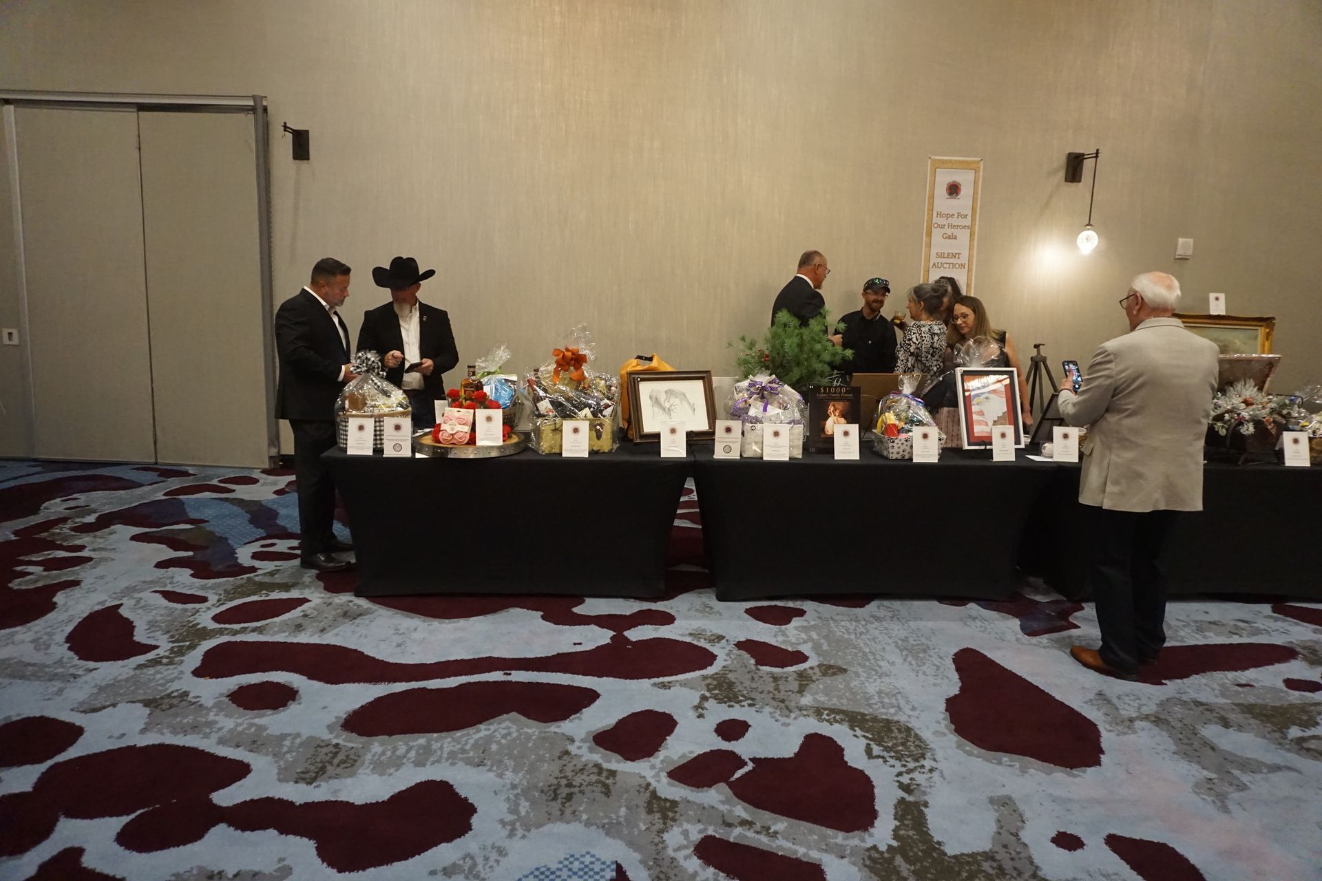 People gather around a long table with gift baskets and auction items in a hotel conference room with patterned carpet.