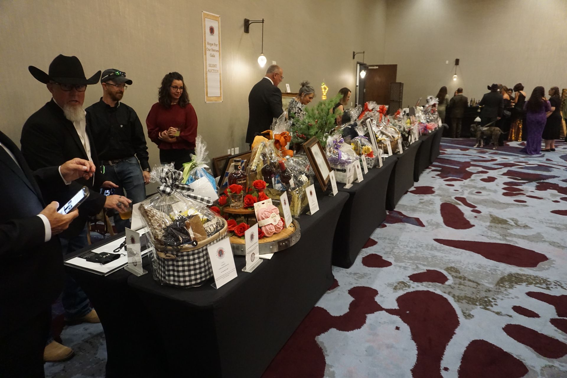 Attendees browse a silent auction table displaying gift baskets at an indoor event with patterned carpeting.