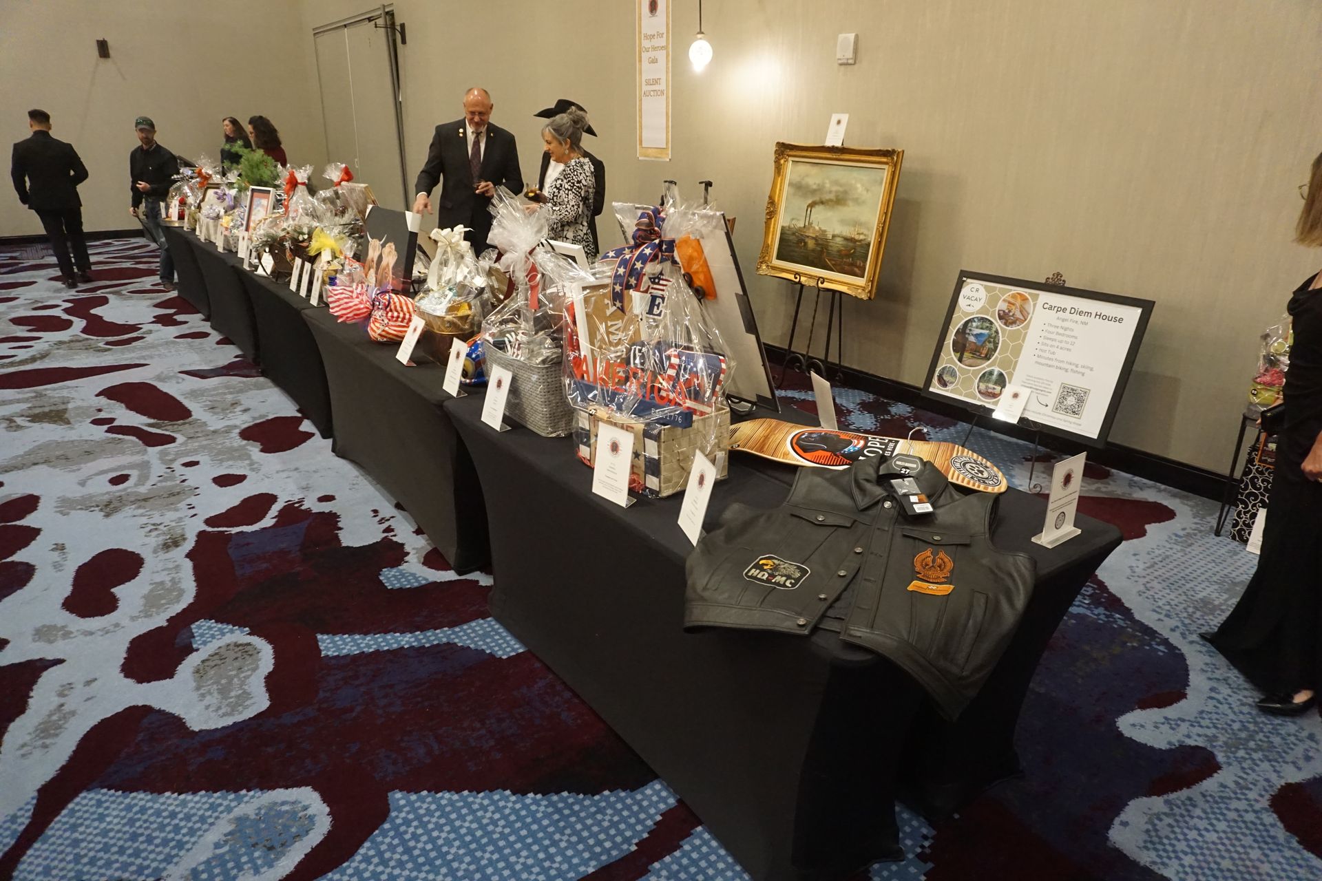 Long tables lined with gift baskets and displayed items for a silent auction, inside a carpeted event hall.