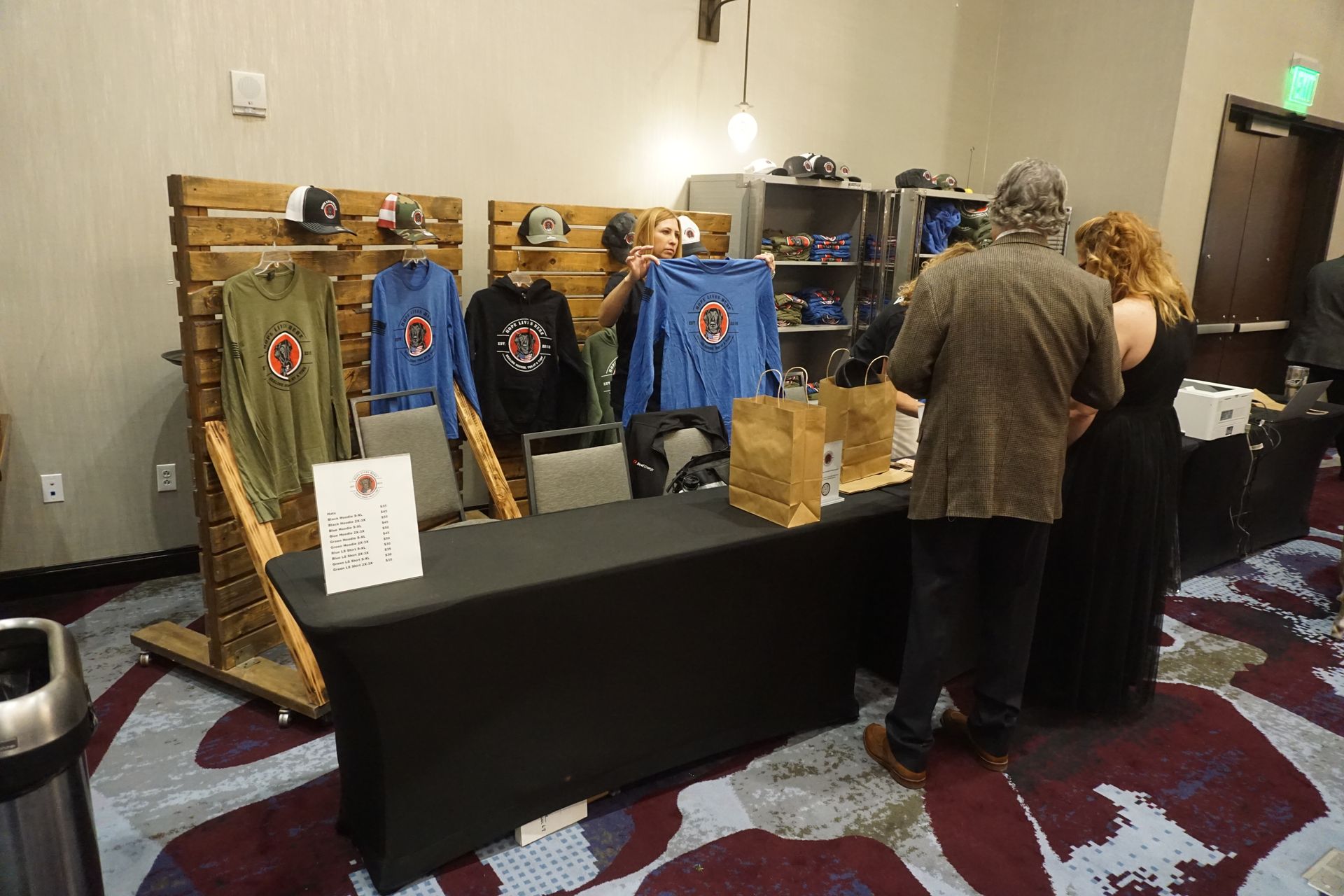 A merch table displays branded shirts and hats on wooden racks as a person helps a visitor inside a conference room.