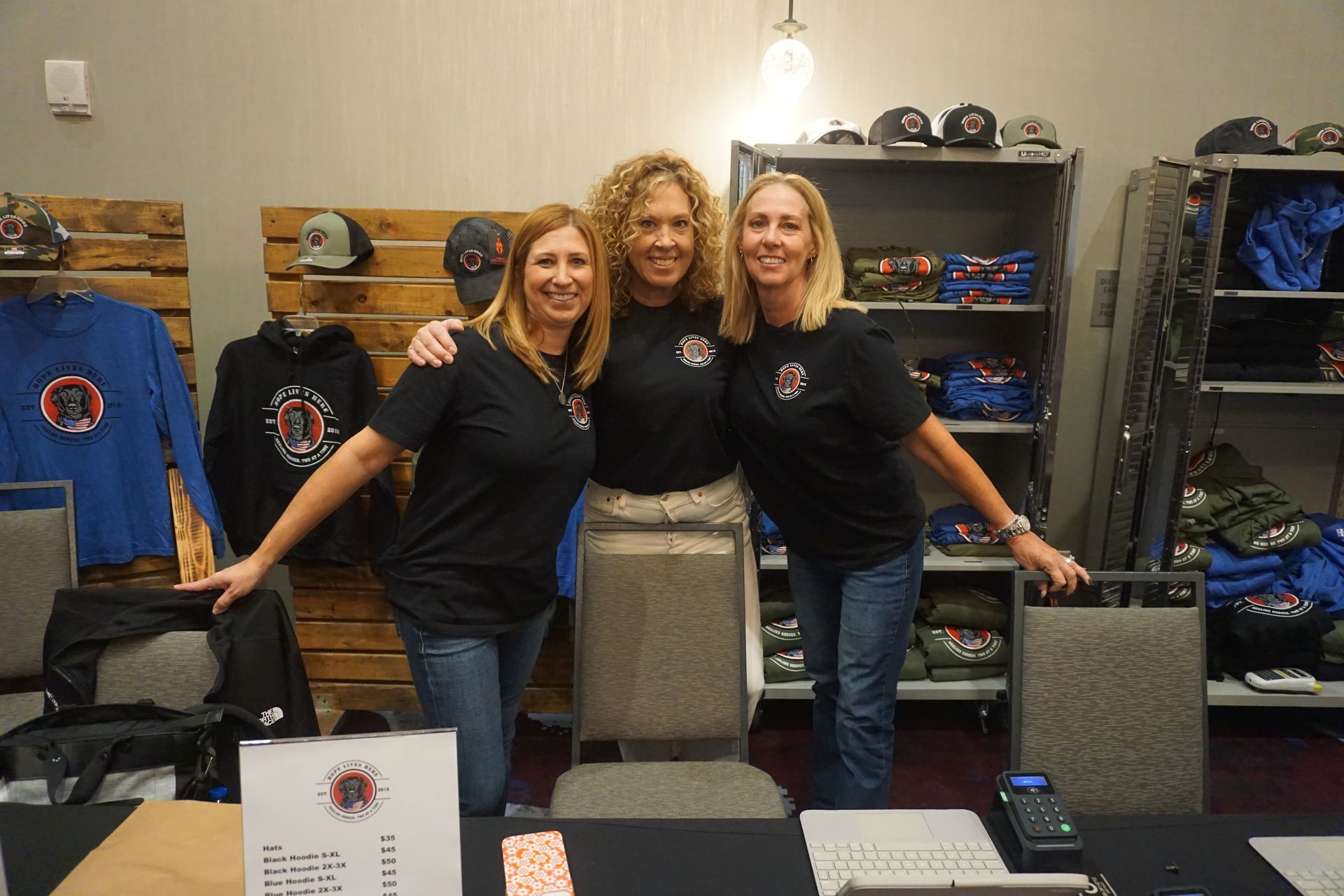 Three people stand behind a merchandise display table in an indoor setting, smiling while wearing black branded shirts.