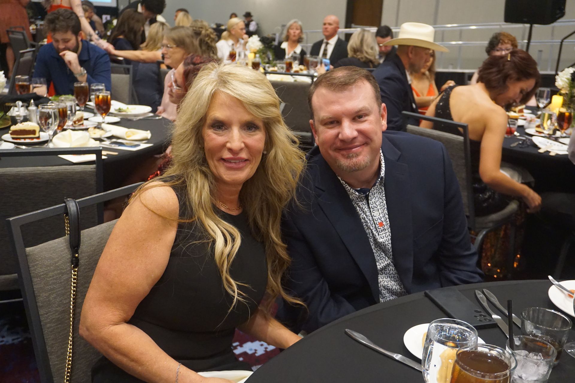 A man and woman pose for a photo at a table in a banquet hall, both smiling at the camera.